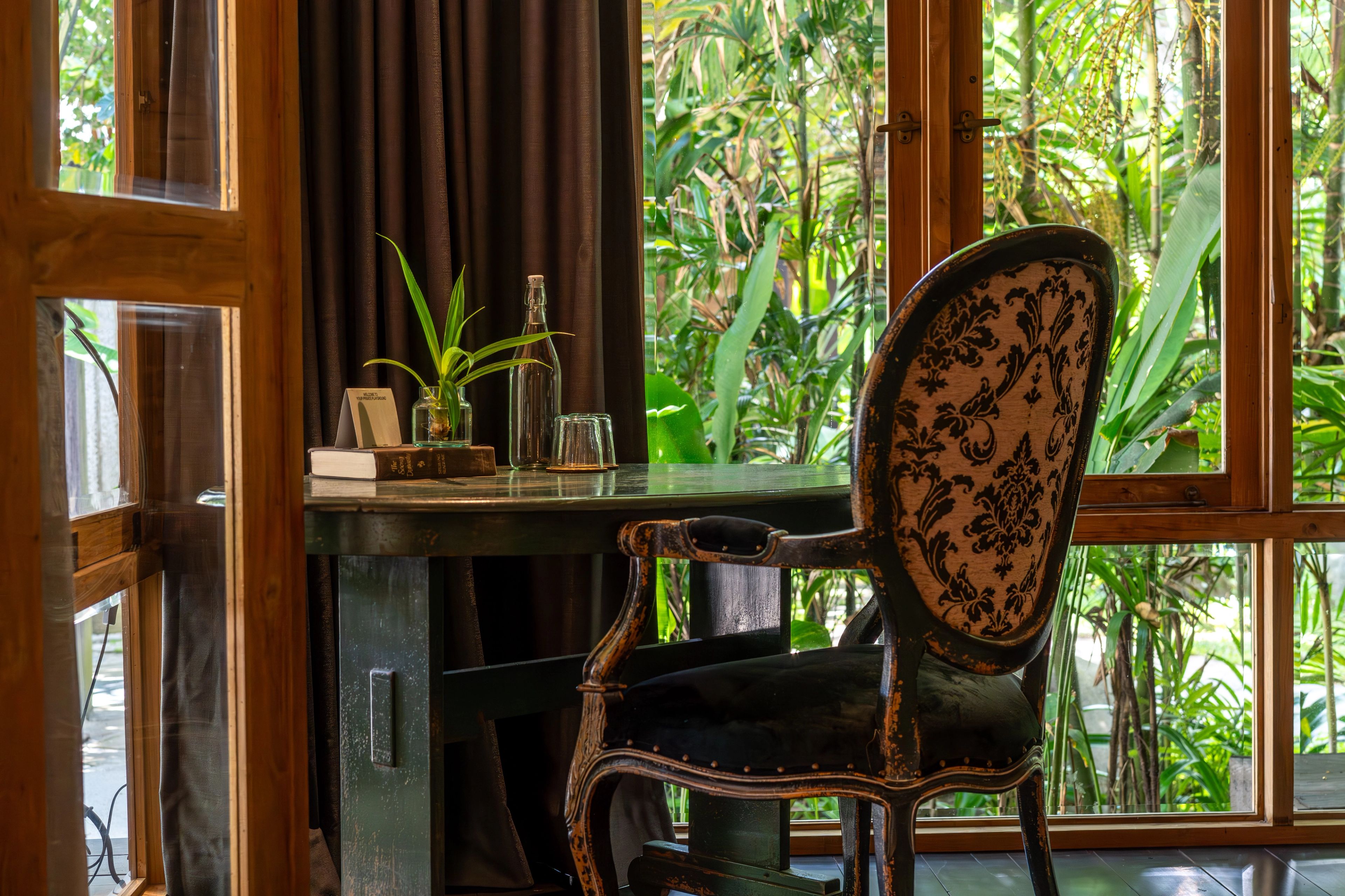 A carved chair at a desk beside tall windows, with planting close outside the glass.