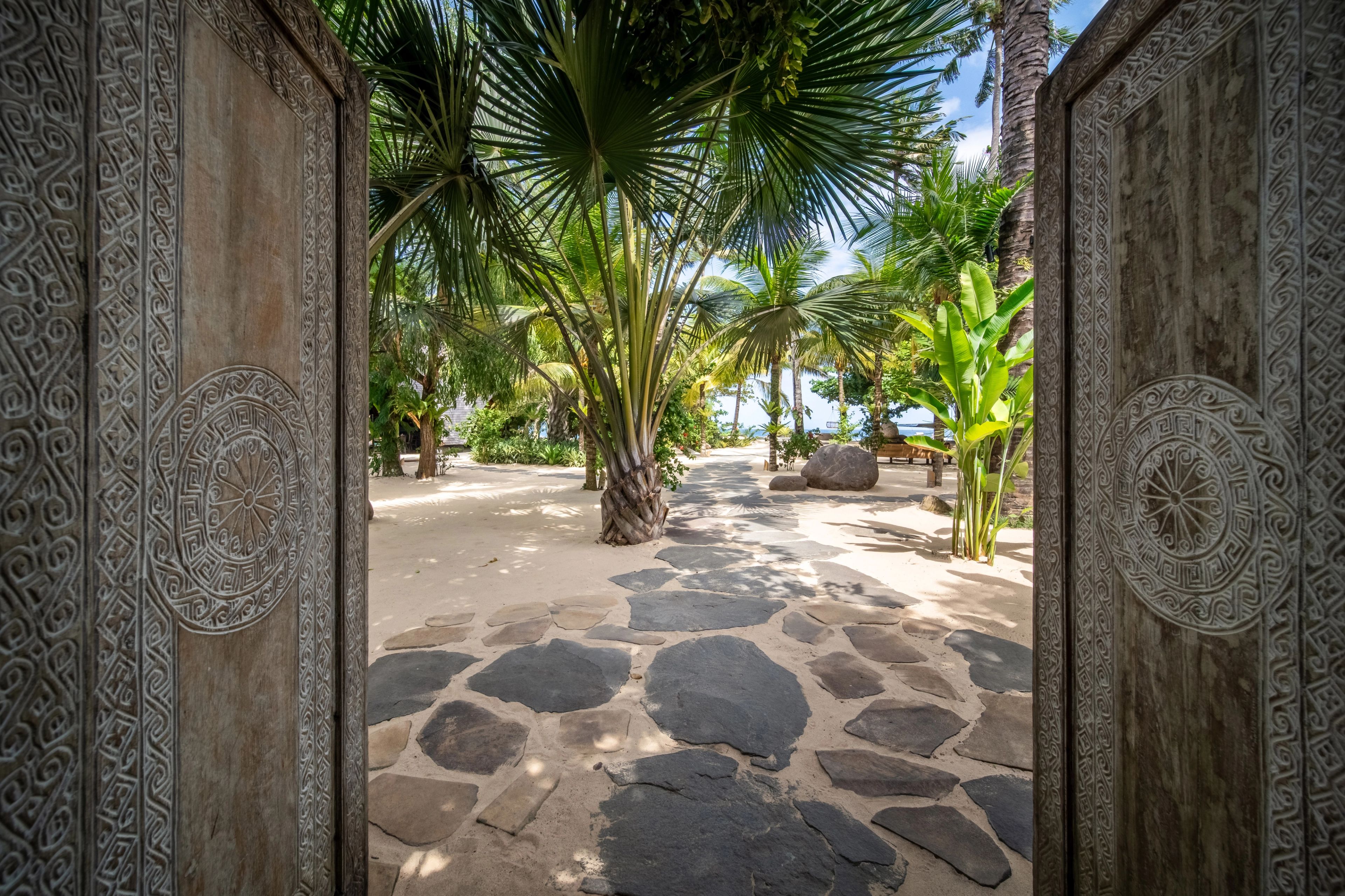 Carved wooden entrance doors set along a tropical garden pathway.