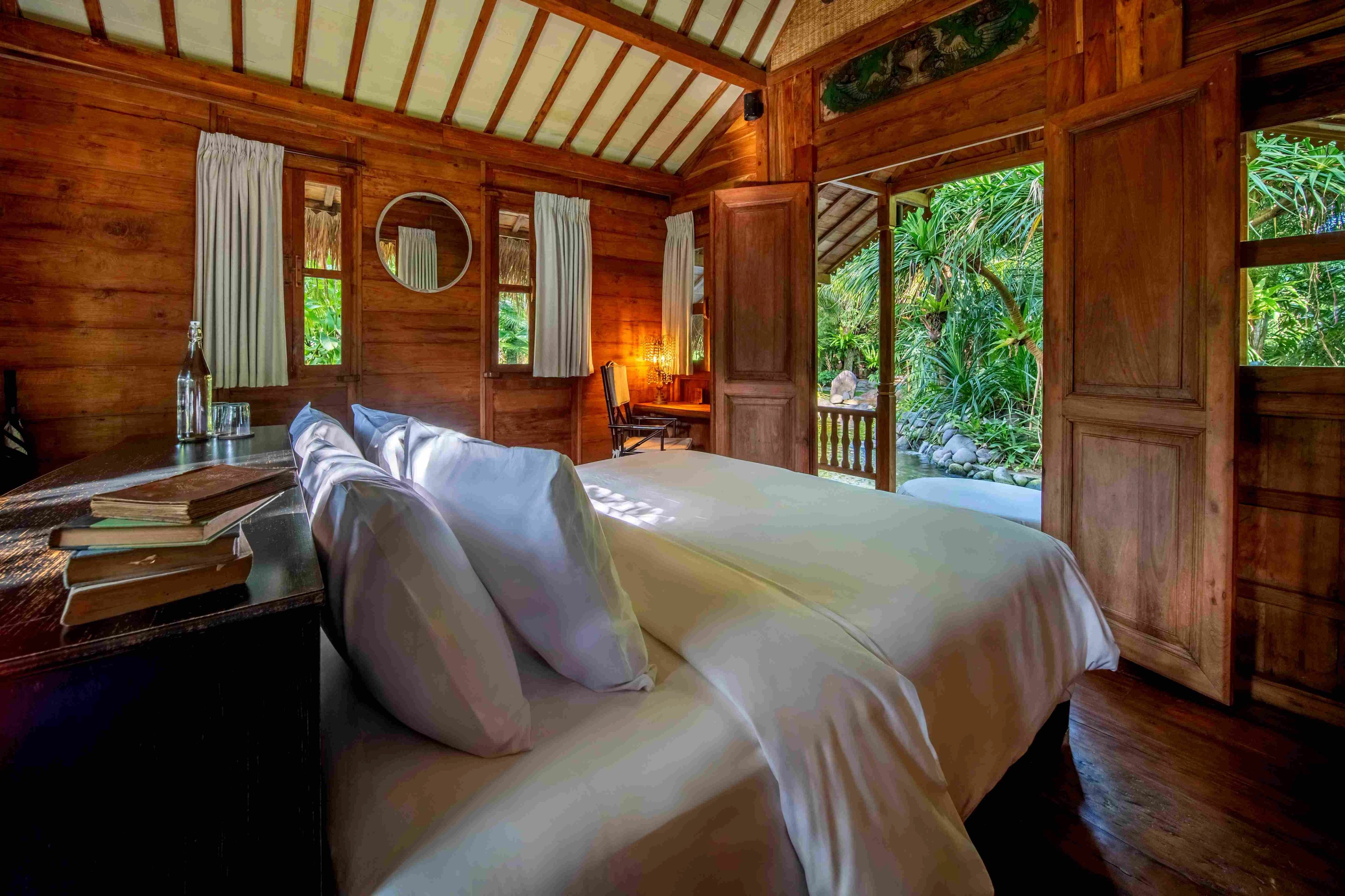 A made bed piled with white pillows in a timber-lined room, with shuttered doors open to the garden.