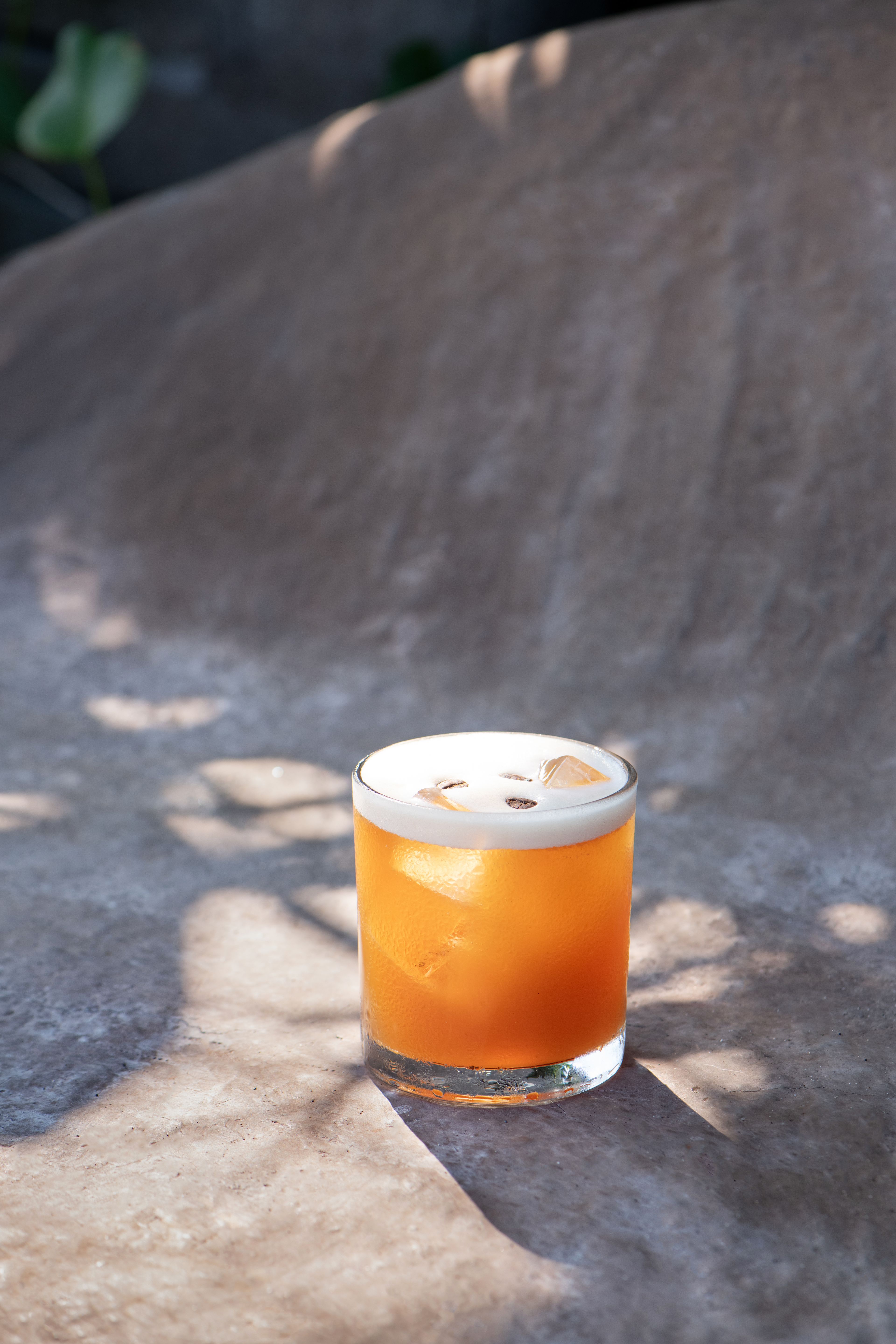 An orange sour topped with foam in a tumbler, dappled light across the wall behind.