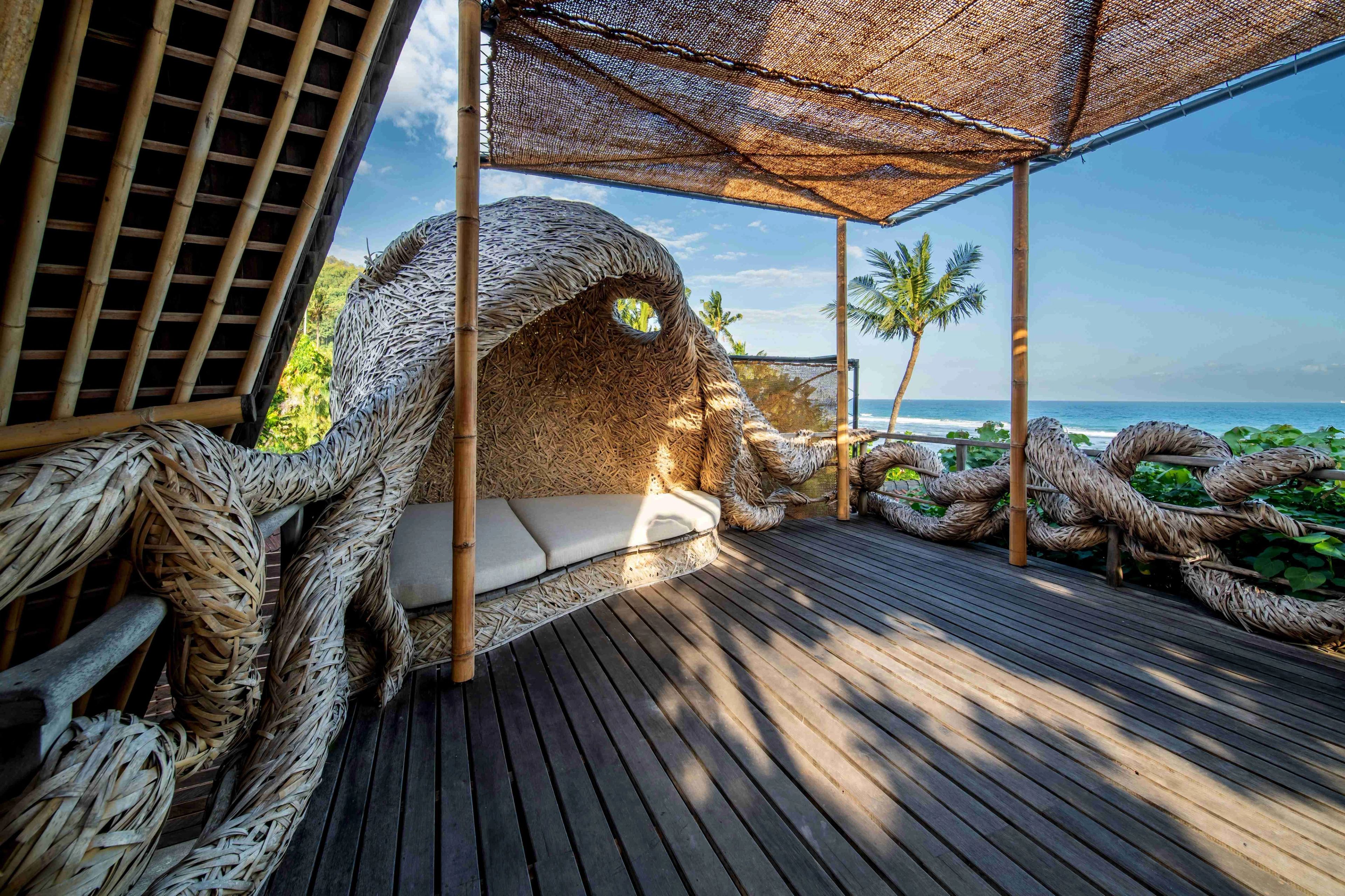 A cushioned bench set into a sculpted woven alcove on the shala deck, with rope forms coiled along the rail and the sea beyond.