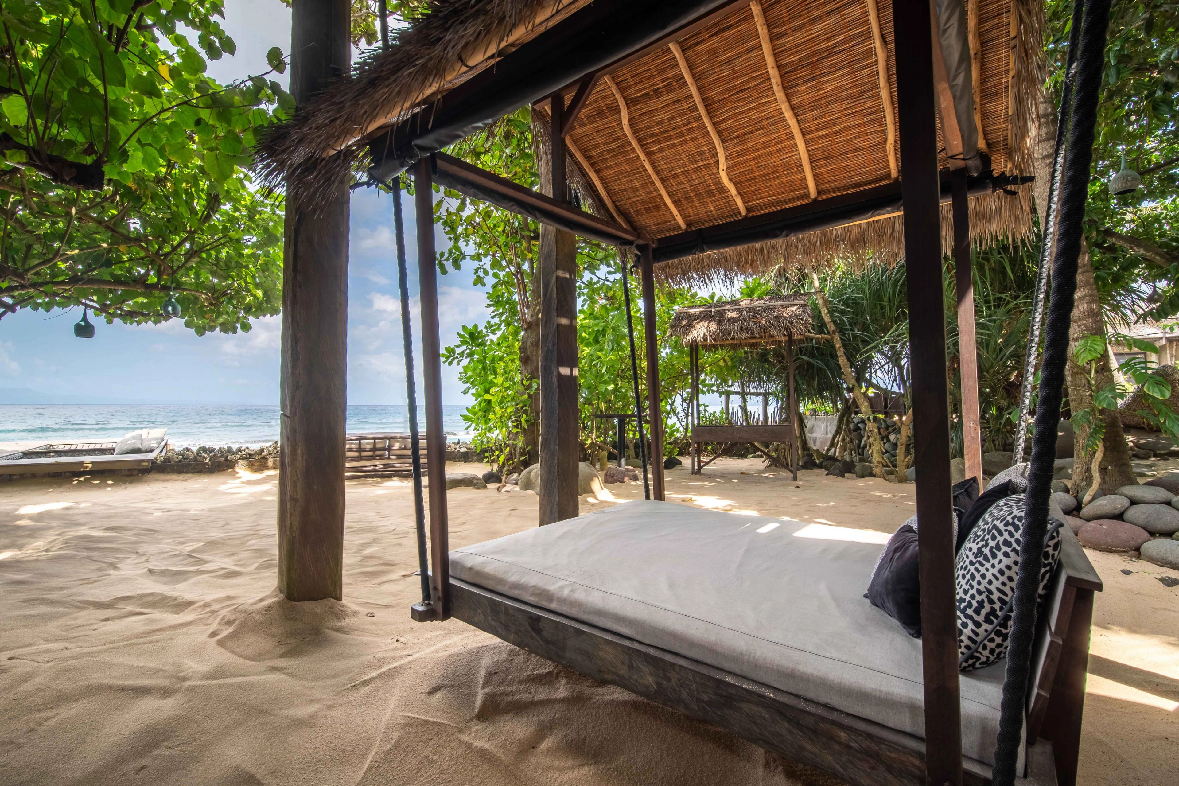 A thatched cabana on the sand holding a cushioned daybed, open on all sides to the sea.
