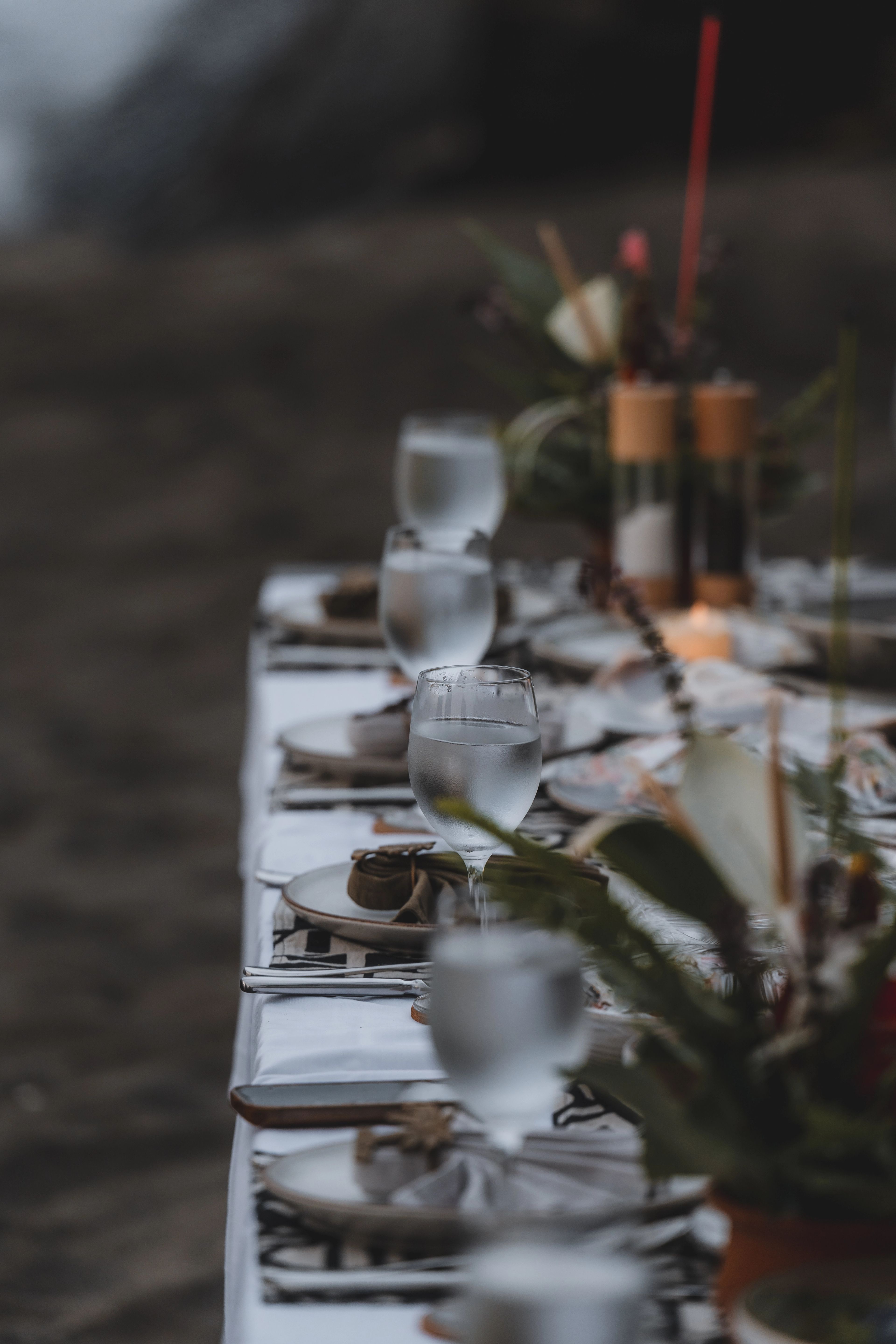 Water glasses, stoneware plates and a run of tropical greenery down a white-clothed table set on the sand at dusk.