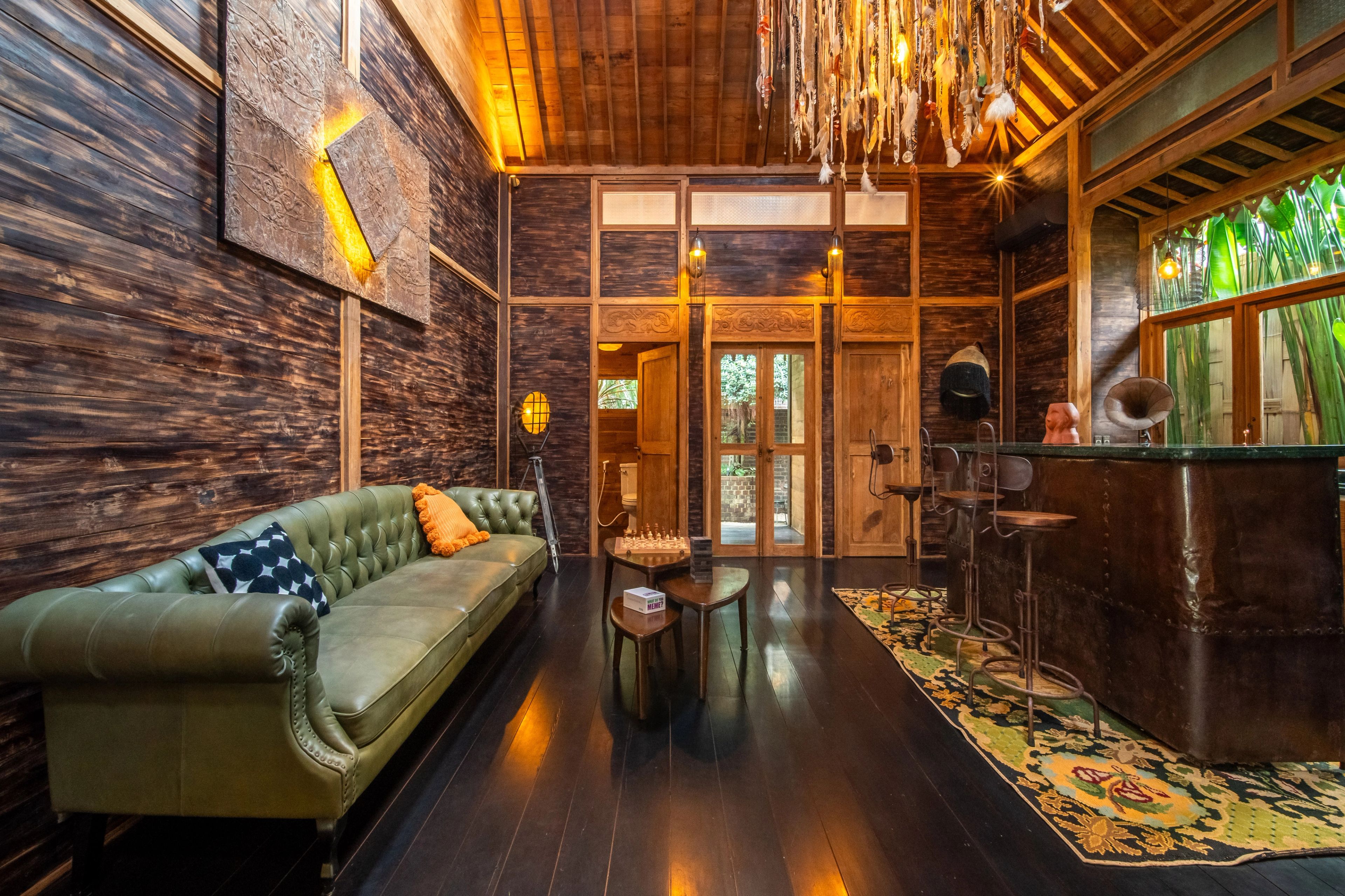 A green leather chesterfield along the dark timber wall, doors open to the garden at the far end.