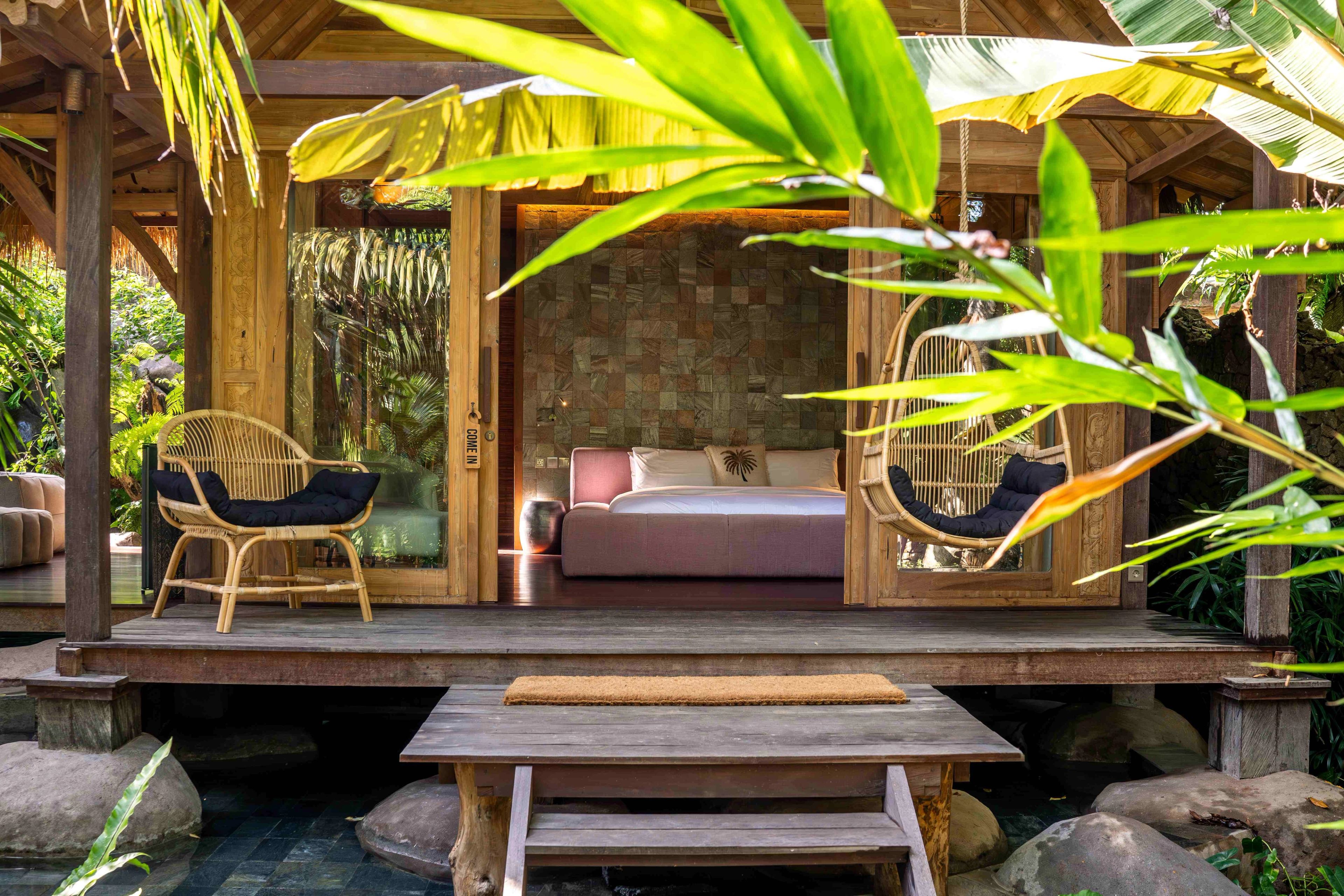 The bedroom seen from the garden through its open doors, a hanging chair and cane armchair on the deck.