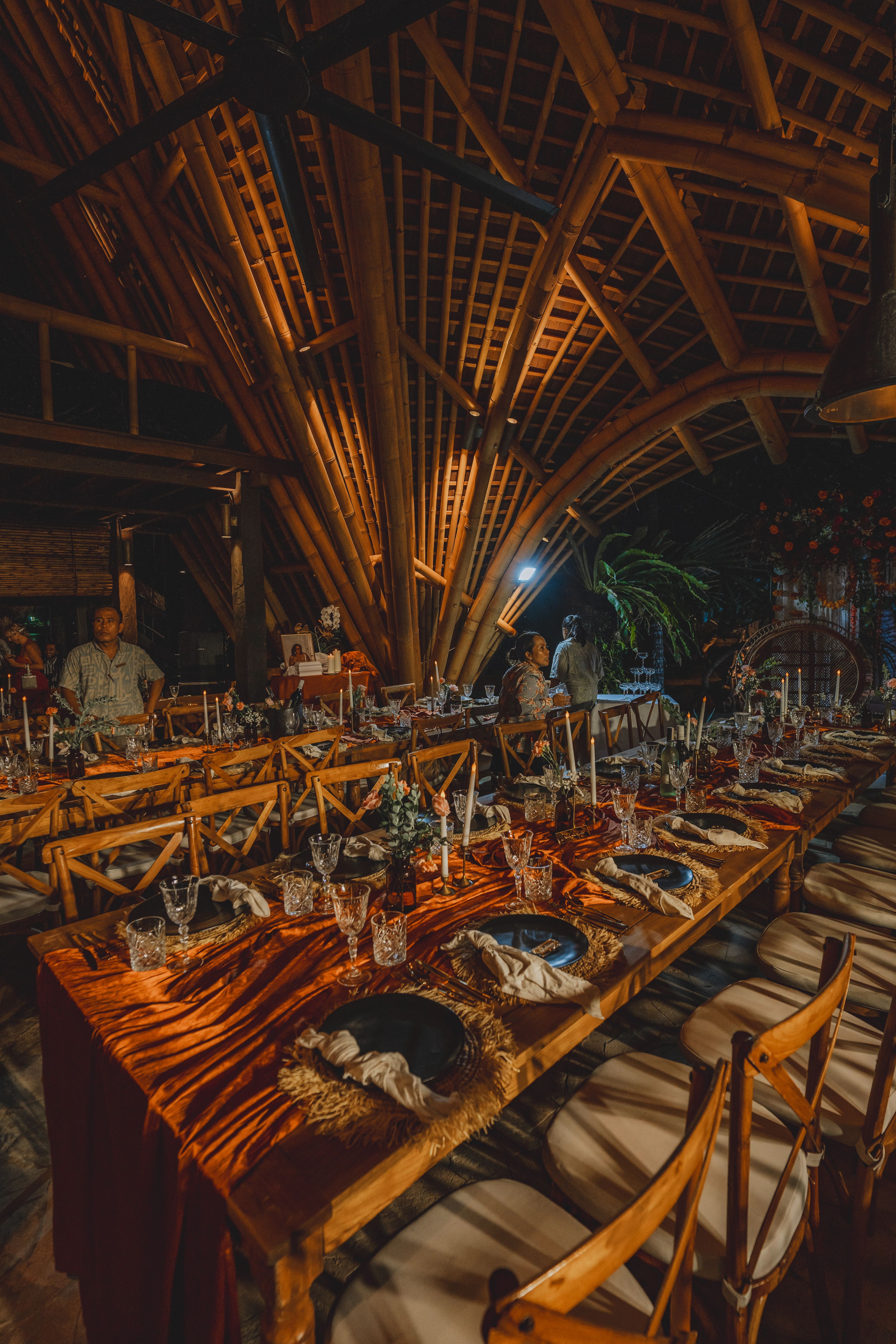 Guests seated at a long table under the bamboo pavilion, candles and glassware down its length.