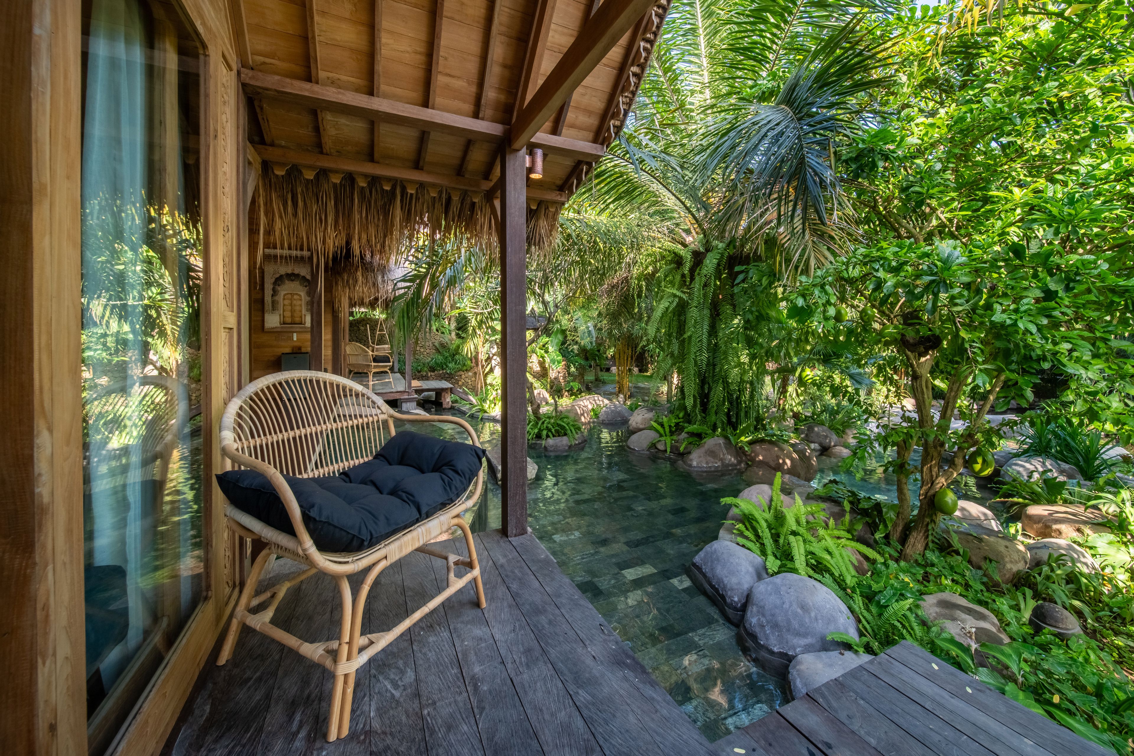 A cane chair with a dark cushion on the villa's deck beside the sliding doors, ferns and boulders beyond.
