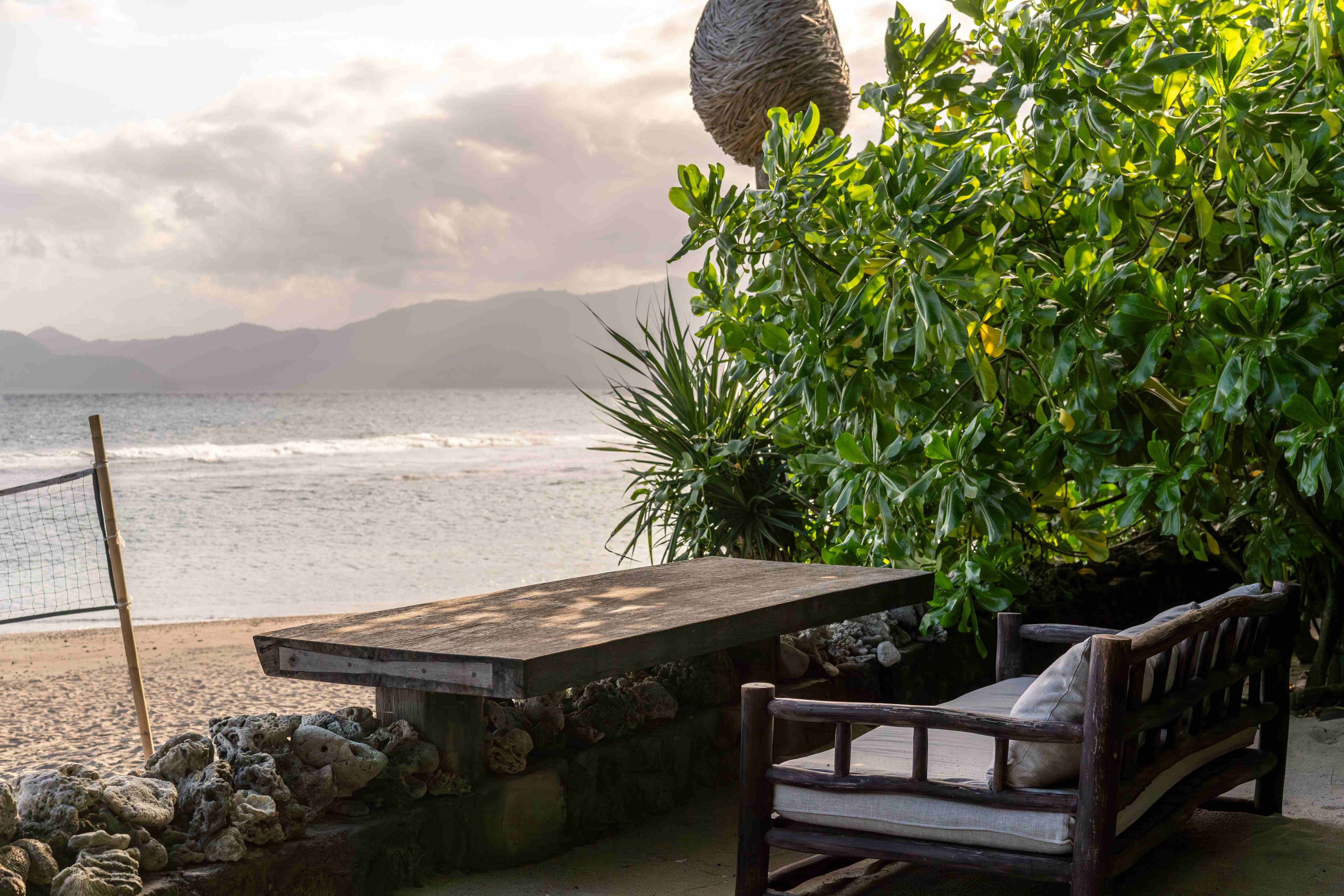 A timber bench and low table on the terrace at dusk, the volleyball net and sea just below.