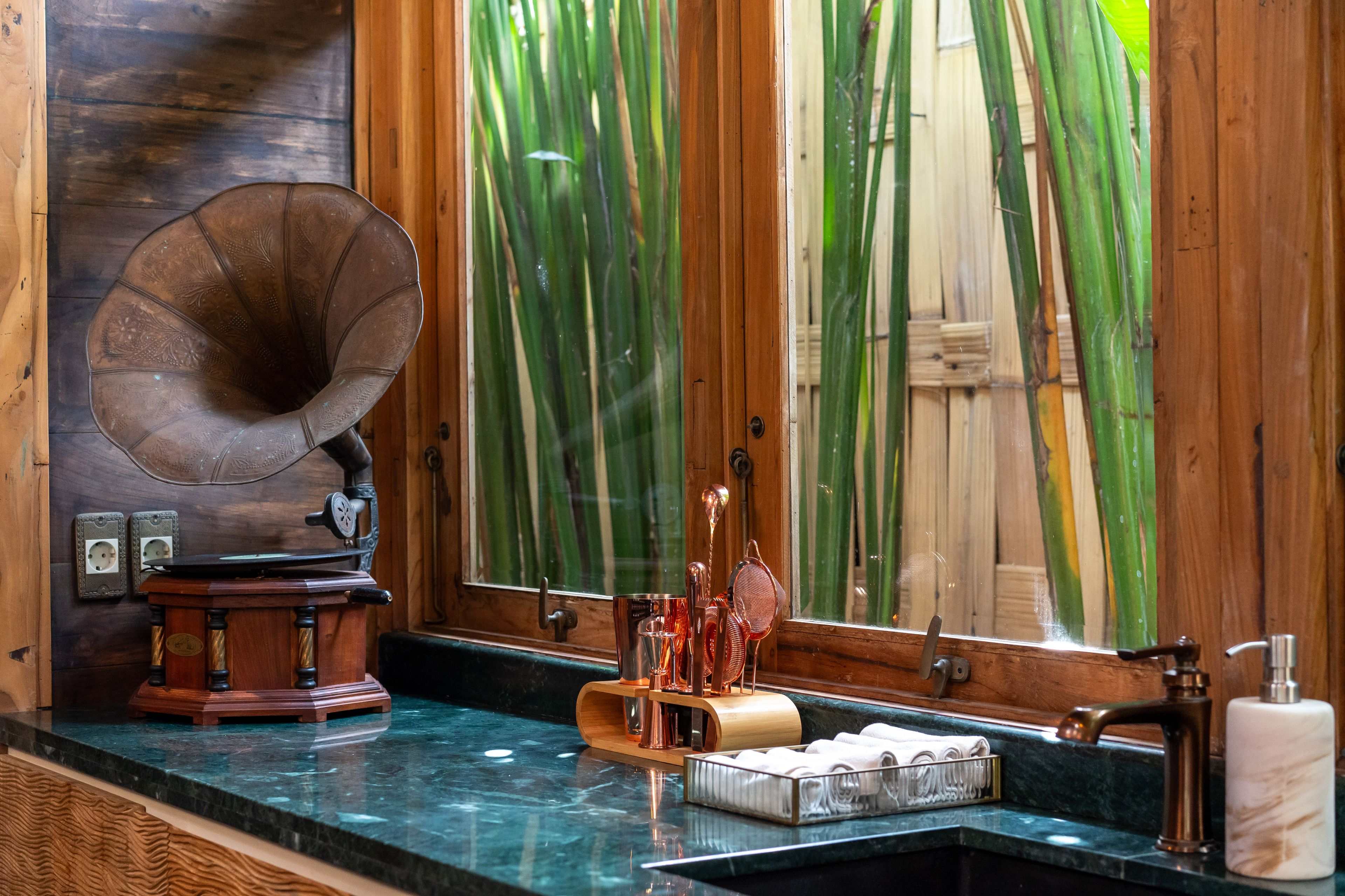 A gramophone and glassware along the green stone bar counter, bamboo growing outside the window.