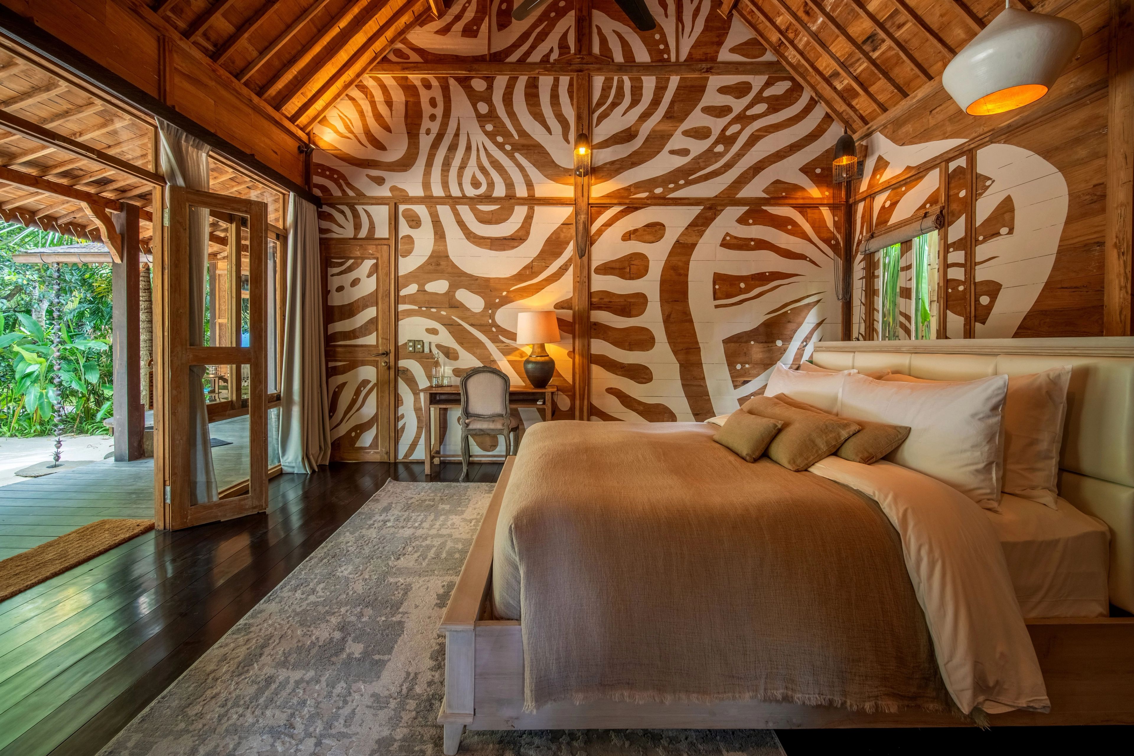 A bed in cream linen against a wall painted with bold cream patterning, doors open to the garden.