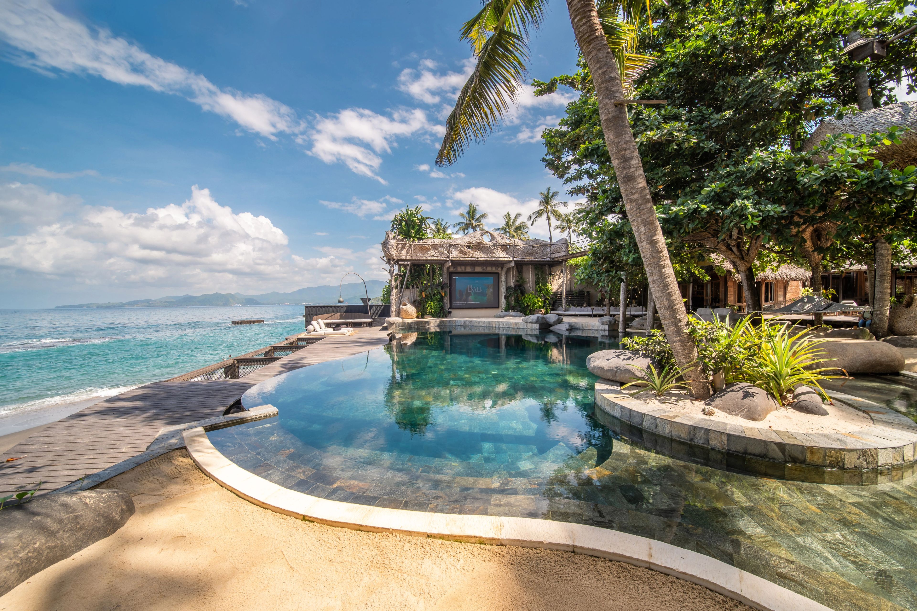 The pool curving past its planted island towards the pavilion and the cinema screen, the sea beyond the far edge.