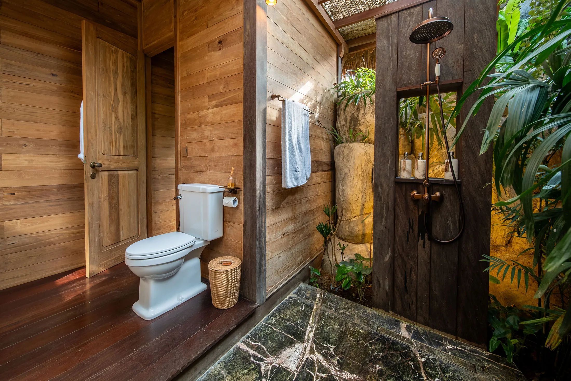 The timber bathroom opening onto a stone-floored outdoor shower with planting all around it.