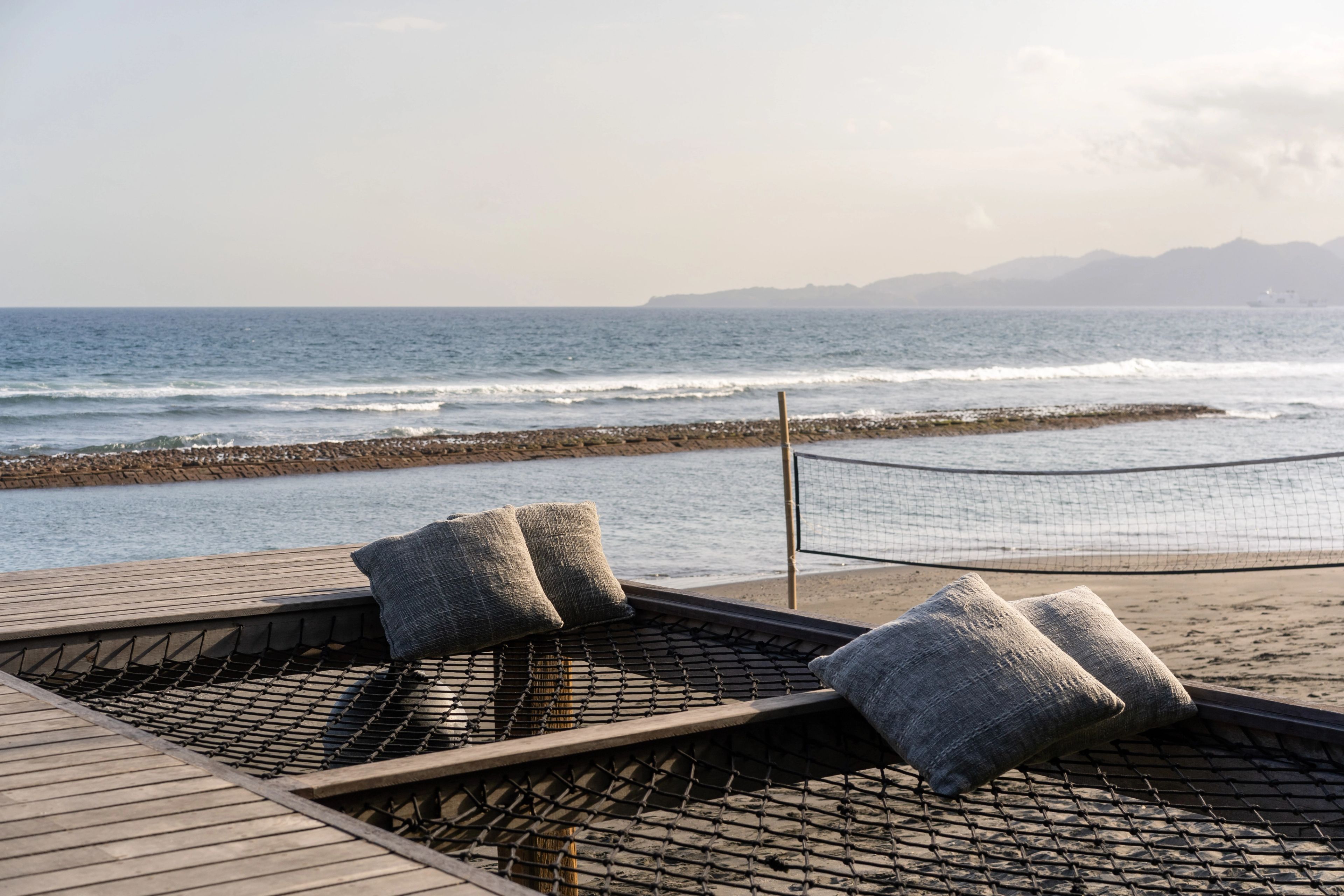 A rope-netted daybed scattered with grey cushions at the deck's edge, above the beach volleyball net and the sea.