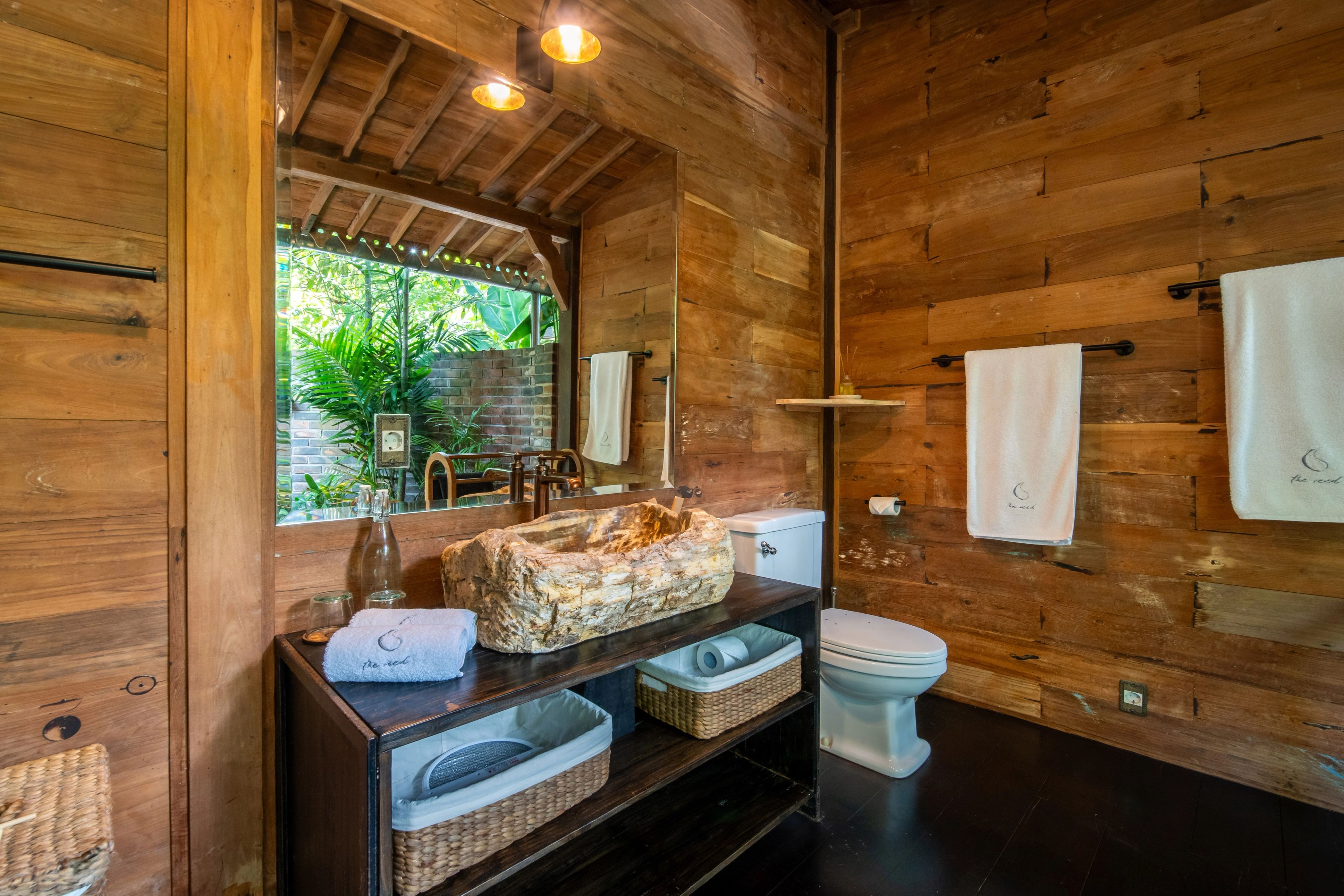 A stone basin on a dark vanity with baskets beneath, a window opening to the planting behind.