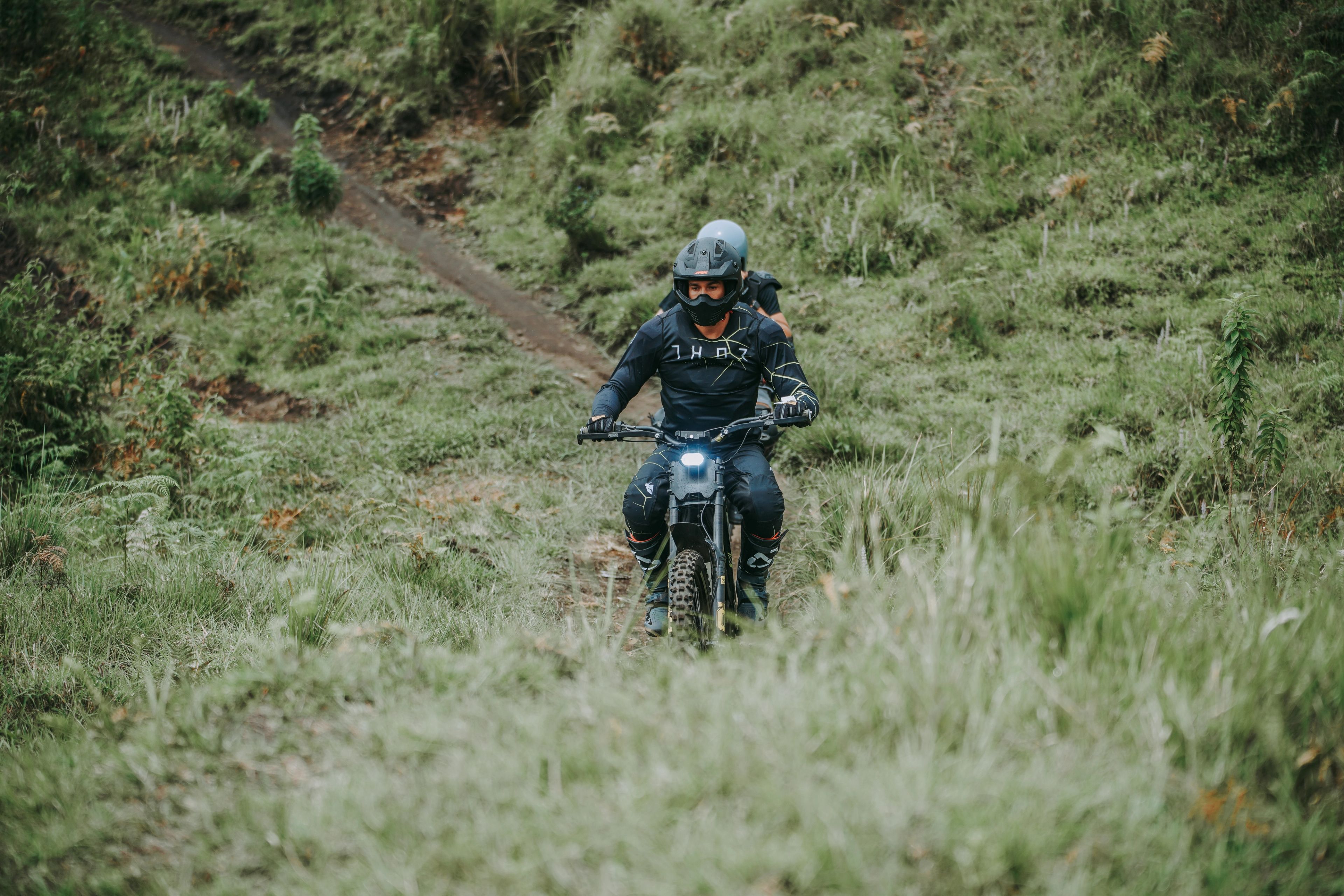 Two riders on electric bikes coming down a green trail between the trees.