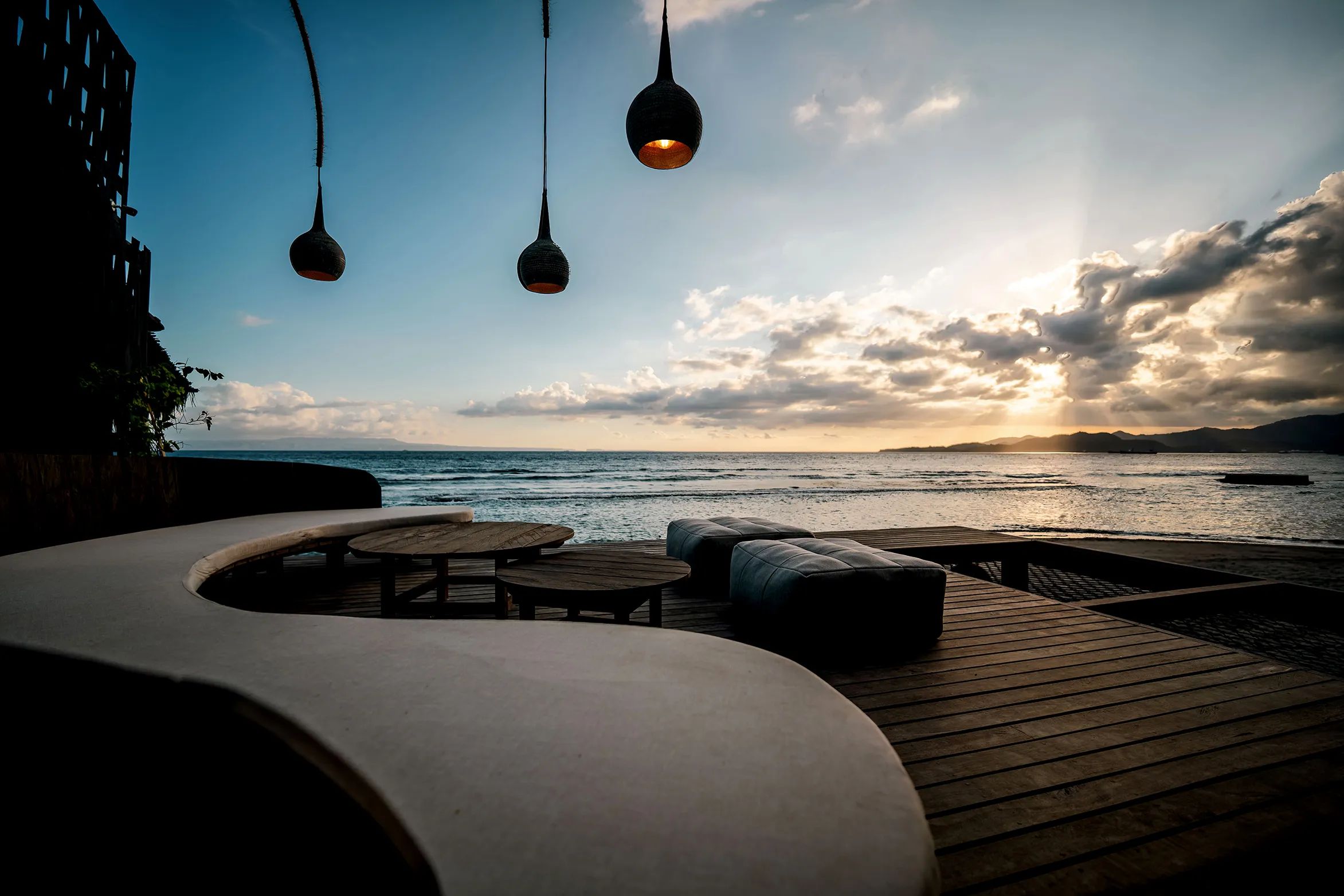 A curved built-in sofa under hanging pendant lamps on the deck, looking out to the sun setting over the sea.