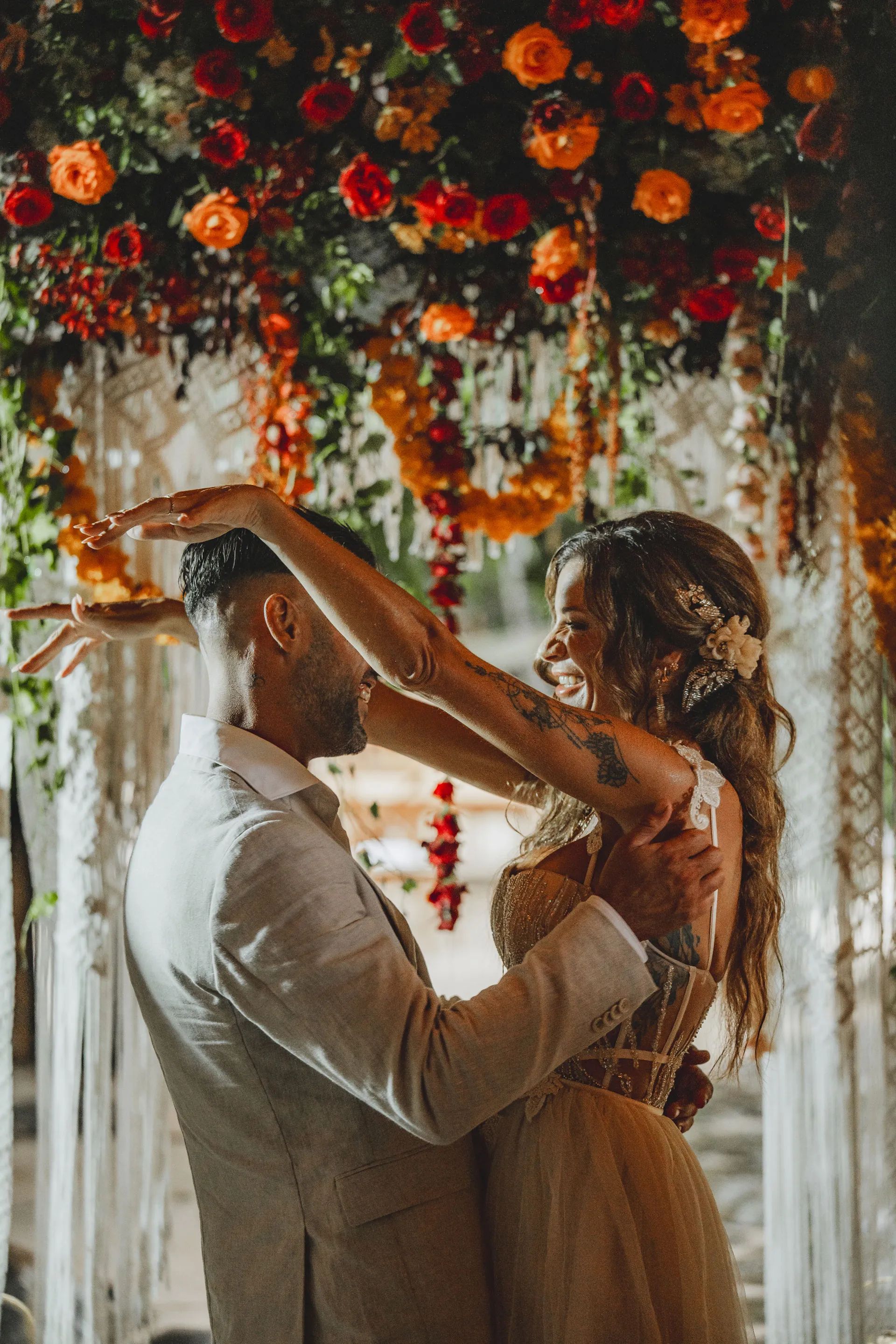 A couple laughing as a garland of marigolds is lifted over them beneath a flowered arch.