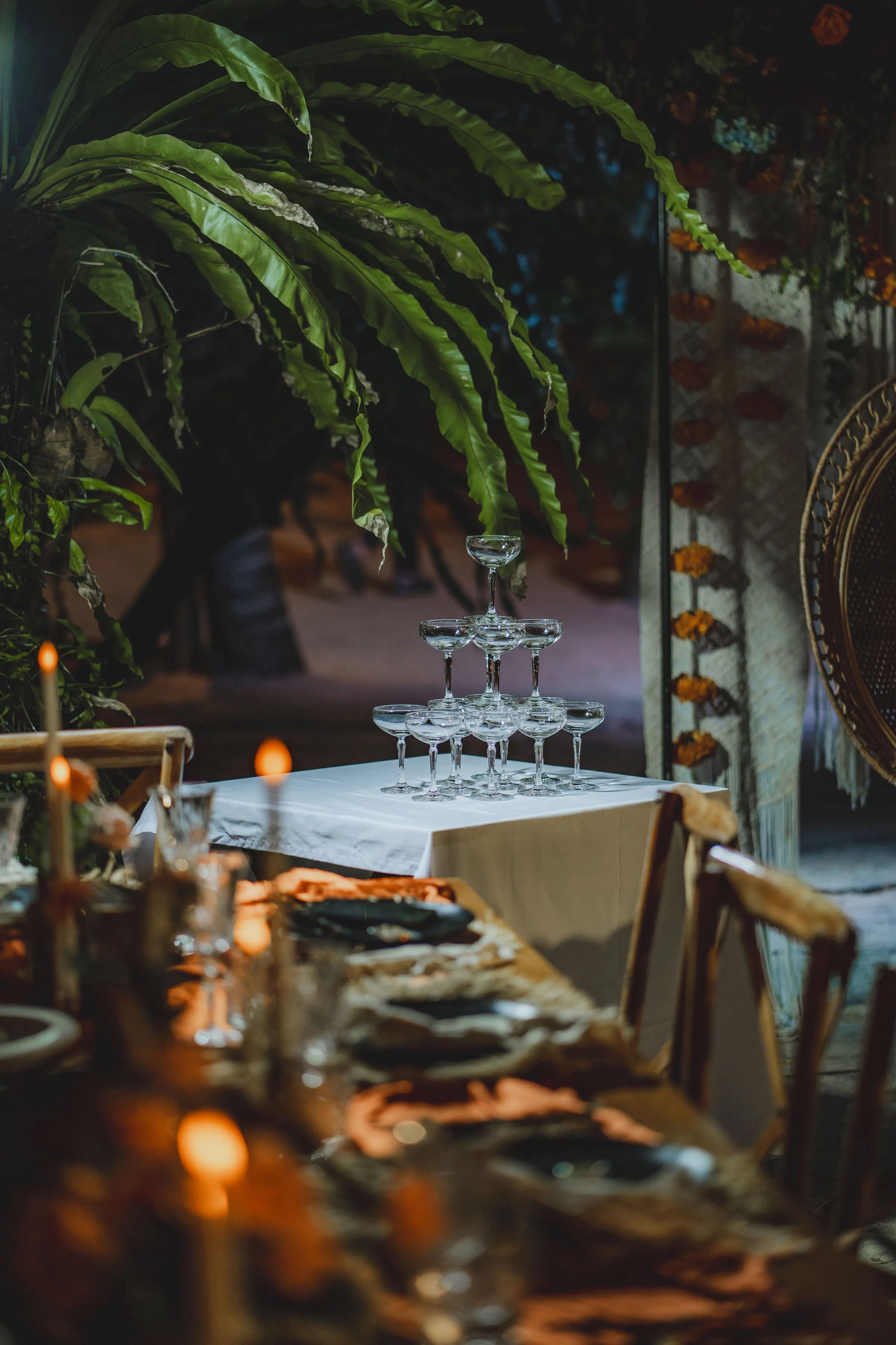 A stacked tower of coupe glasses on a linen-covered table beside a peacock chair, lit by candles.