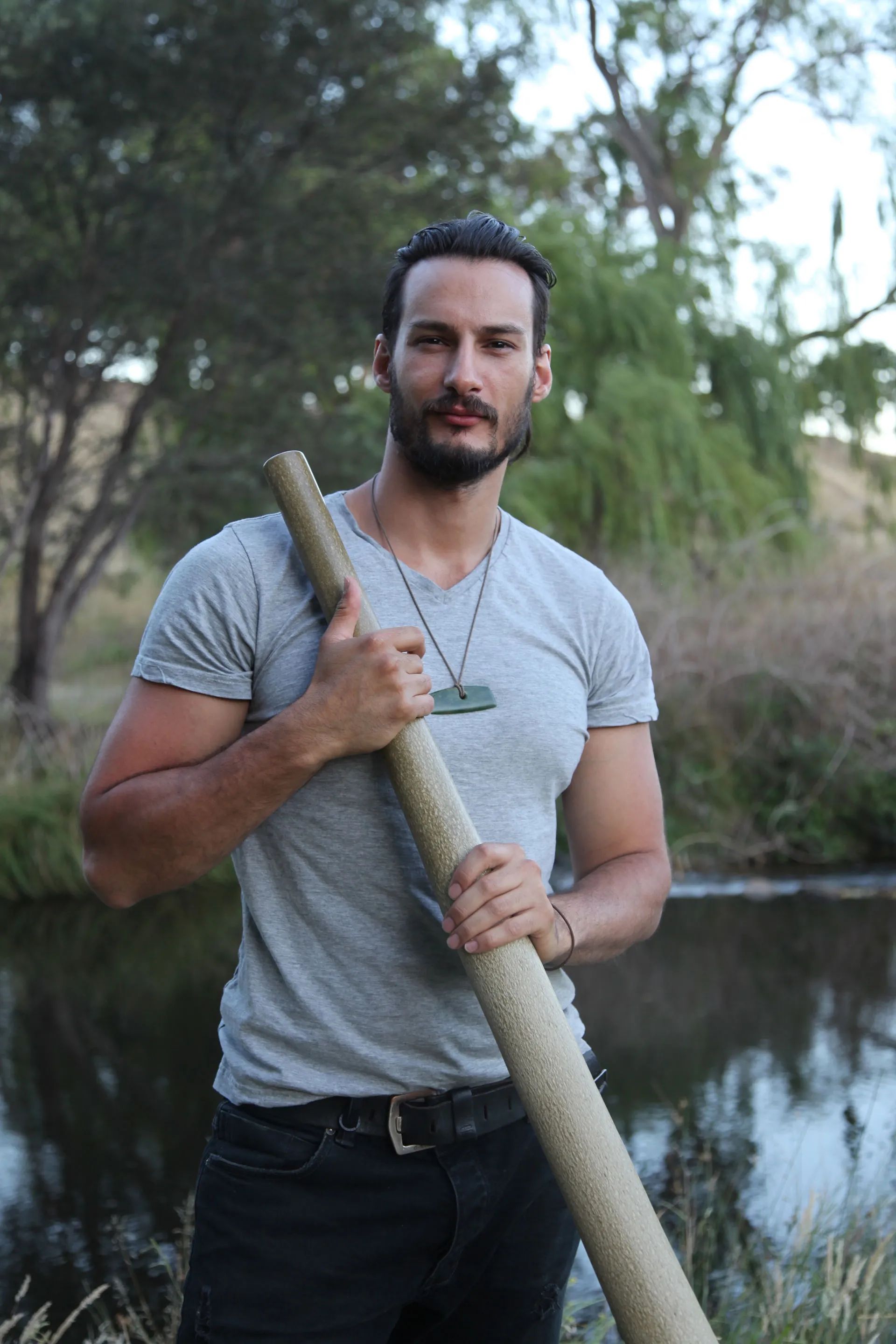 A practitioner holding a wooden rainstick against his chest beside the river.