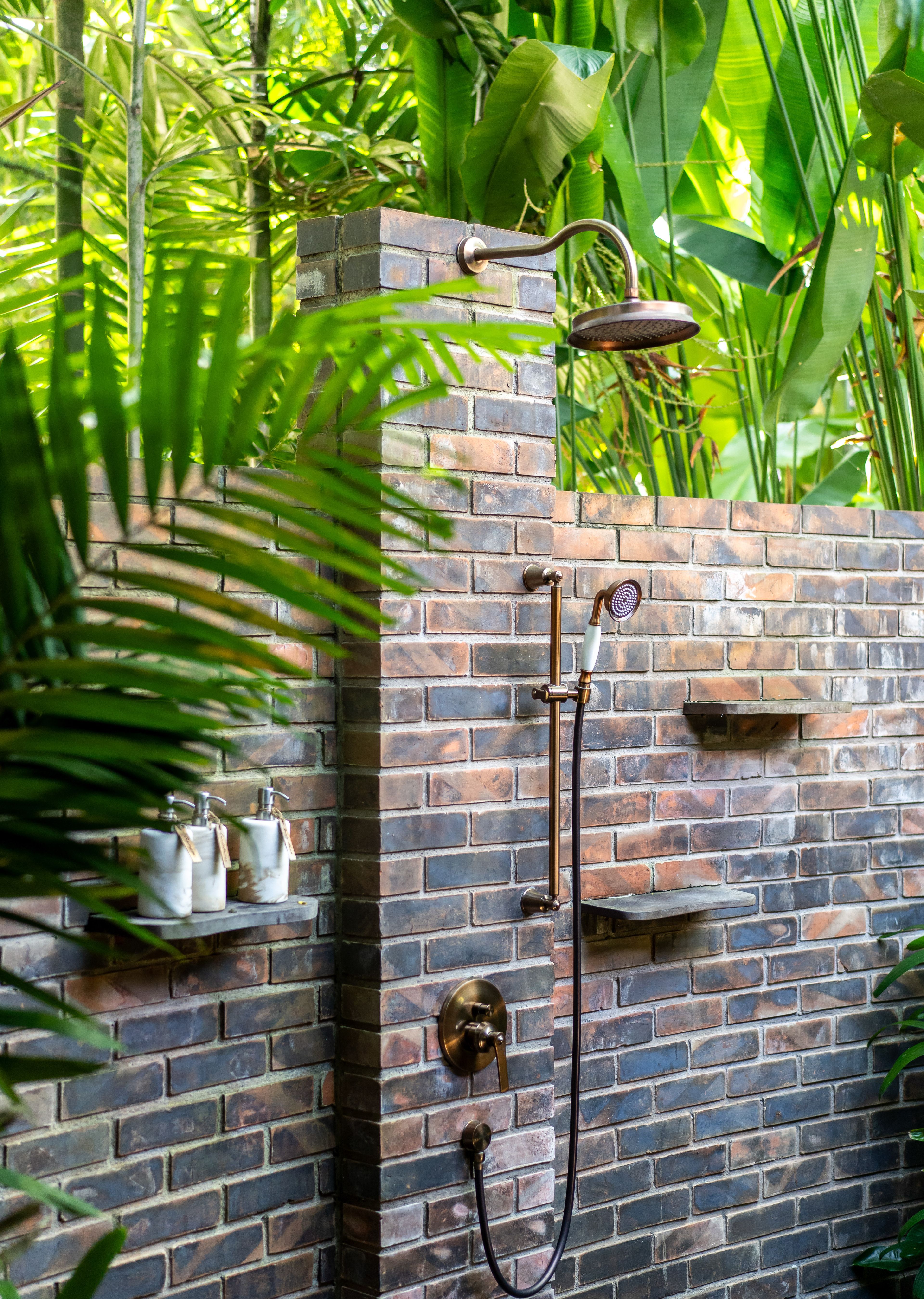 An outdoor shower on a brick wall, amenity bottles on a stone shelf beside it, palms in the foreground.