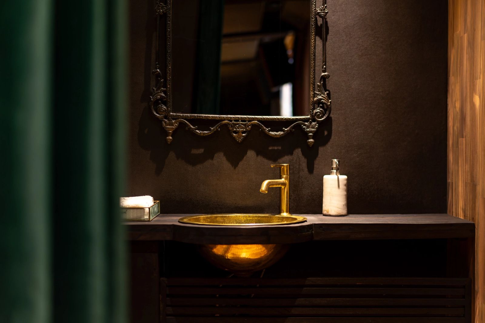 A brass basin and tap under a carved mirror, beside a dark green column in the powder room.