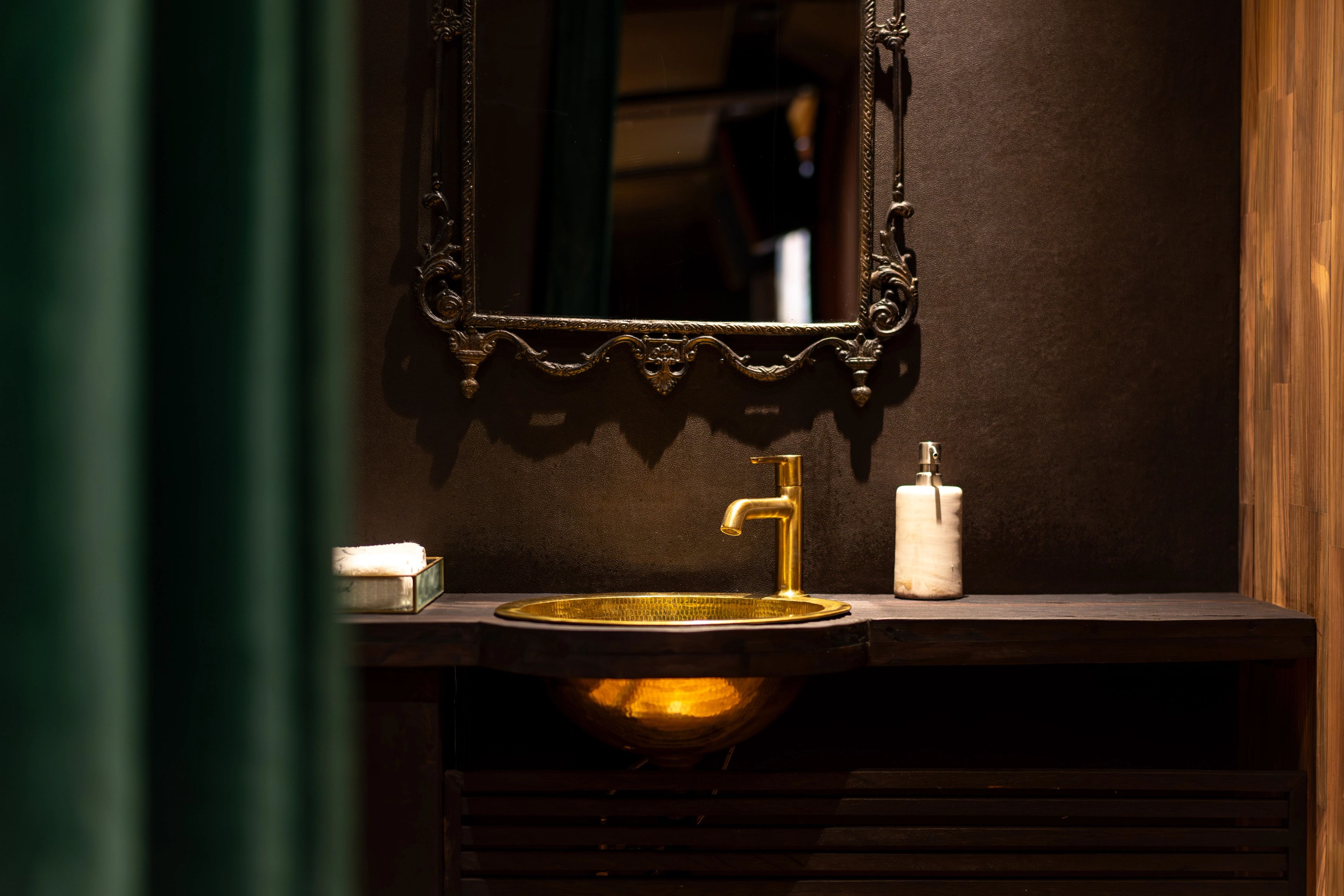 A brass basin and tap under a carved mirror, beside a dark green column in the powder room.