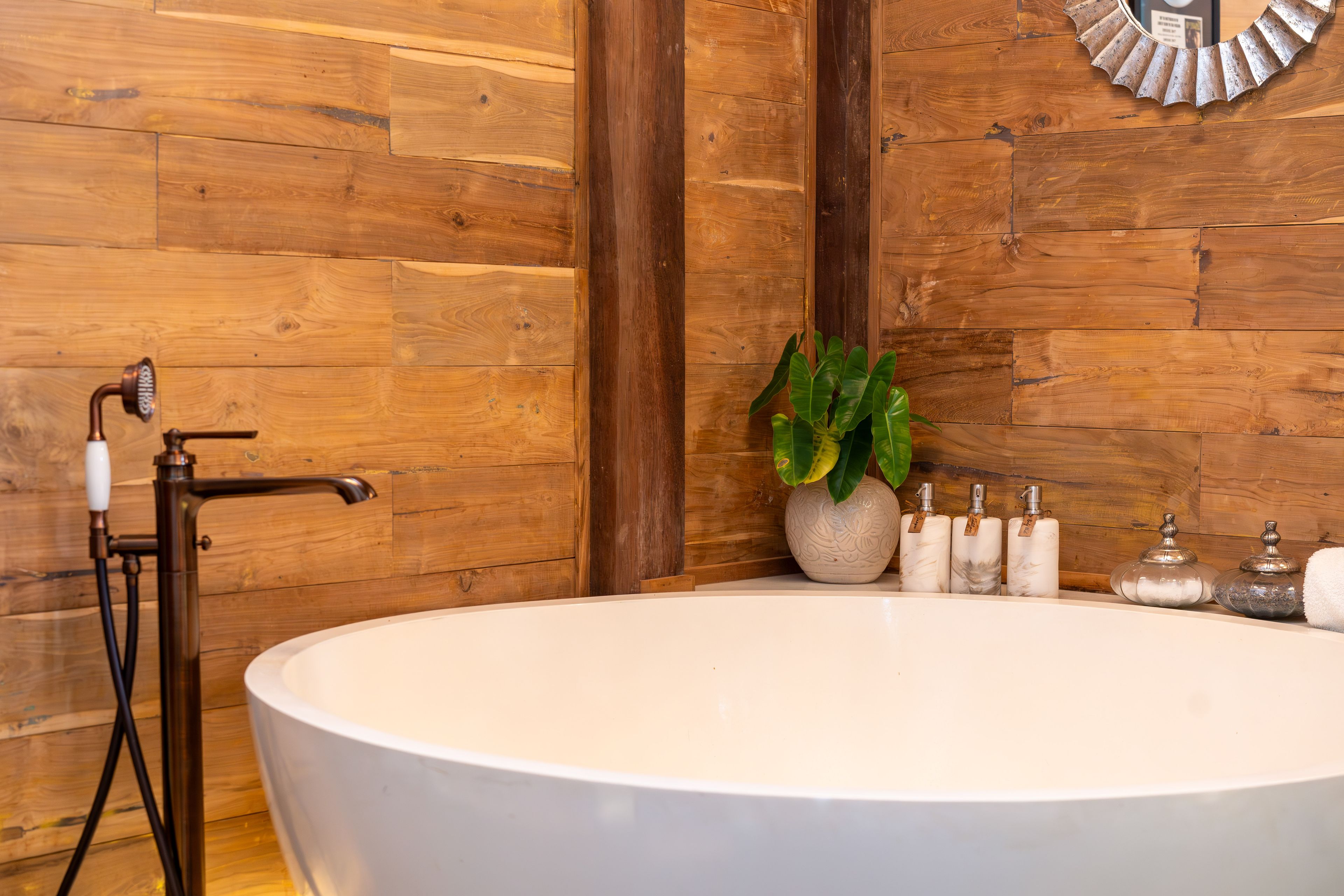 A round freestanding bath against the timber wall, amenities and a plant on the ledge behind.