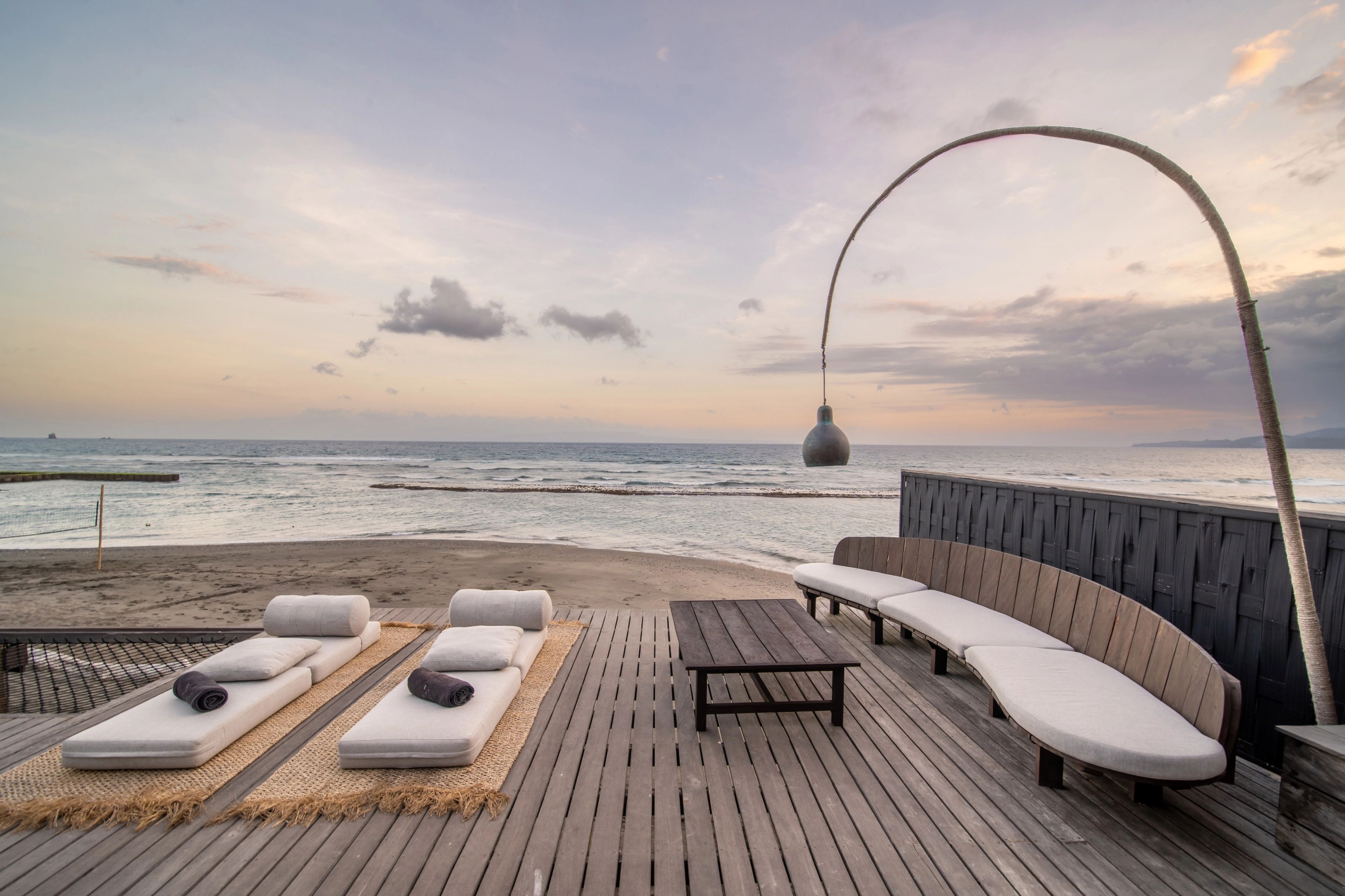 Daybeds and a curved bench on the timber deck under a tall arched lamp, facing the sea at dusk.