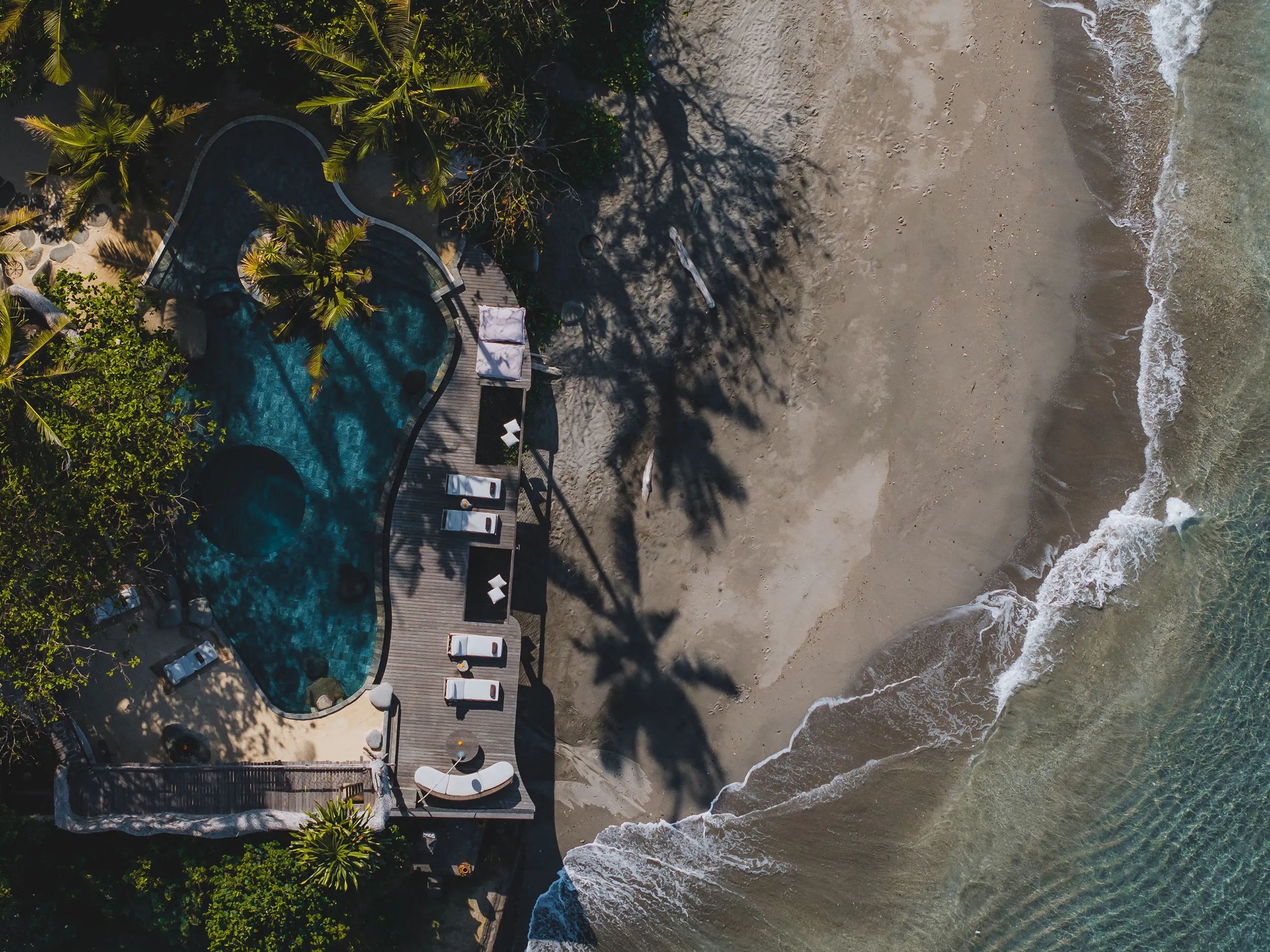 The pool, loungers and palm shadows seen straight down from the air, the surf running along the sand beside it.