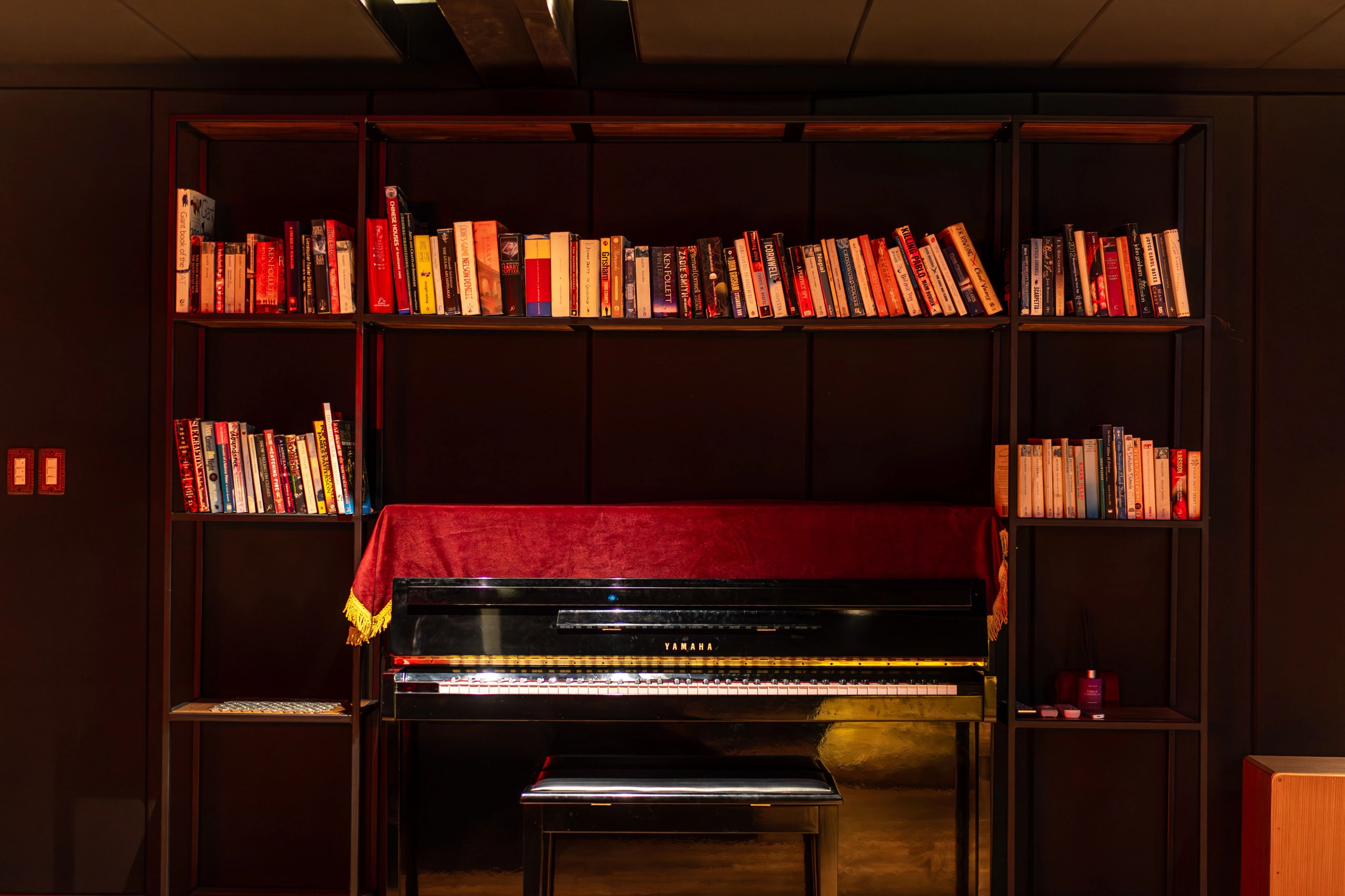 An upright piano set into a full-height bookshelf, its stool tucked beneath and a red runner across the lid.