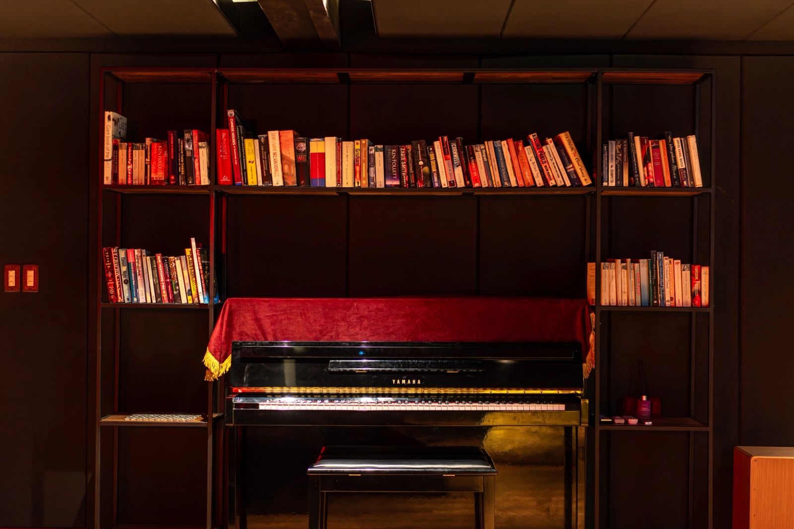 An upright piano set into a full-height bookshelf, its stool tucked beneath and a red runner across the lid.
