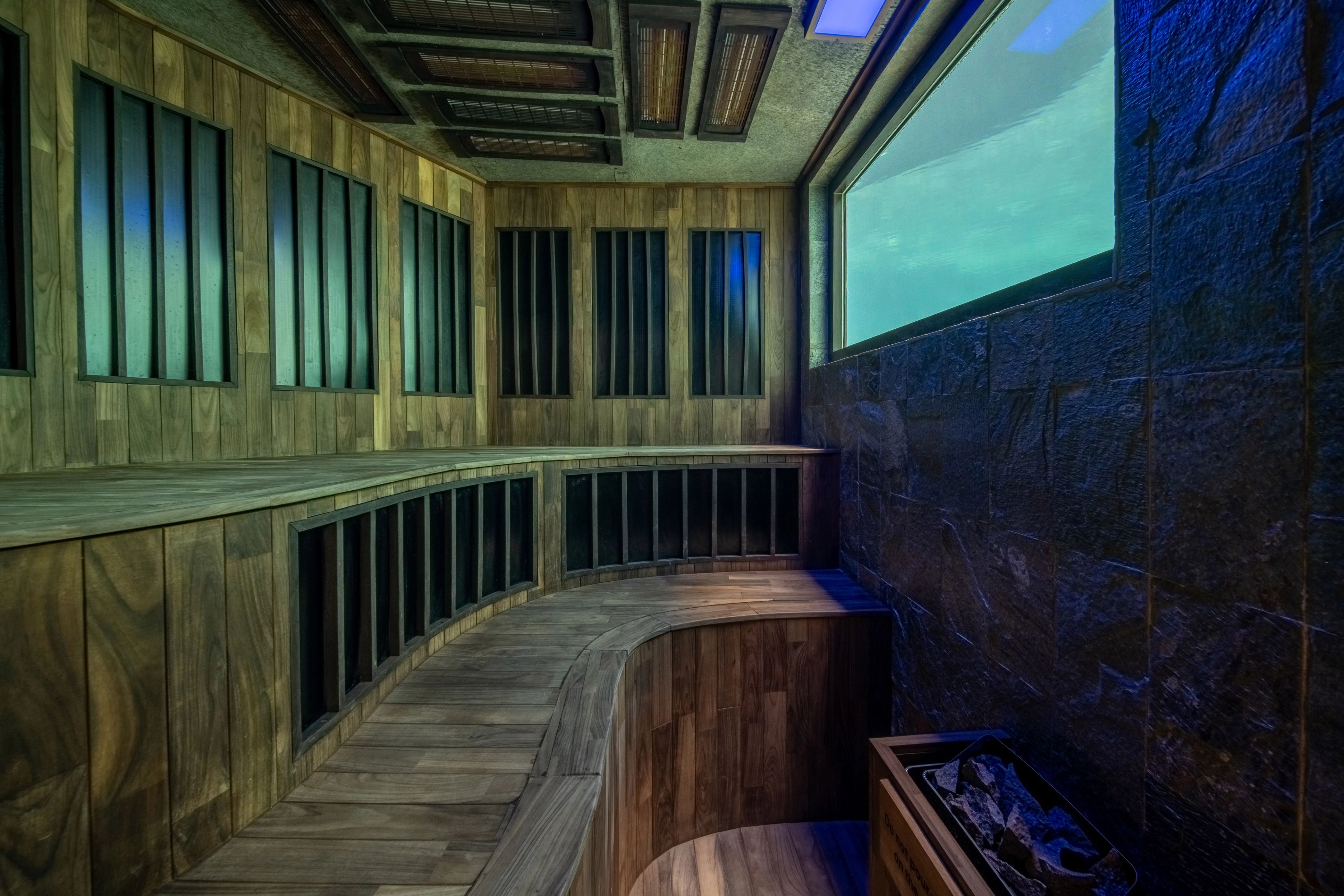 Curved timber sauna benches facing a lit underwater window, the heater at the near end.