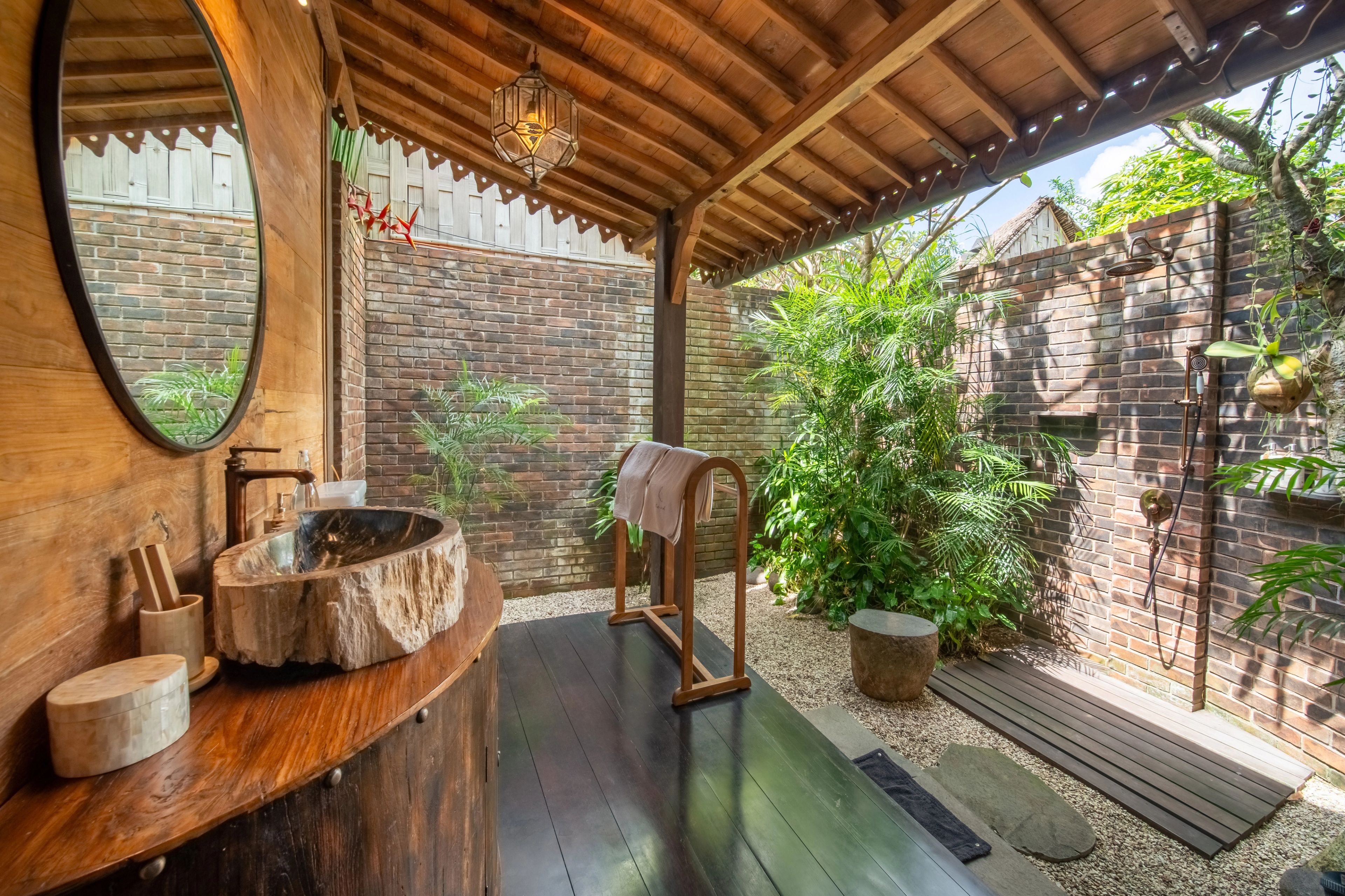 A stone basin on a curved timber vanity under a round mirror, opening onto the walled outdoor bathroom.