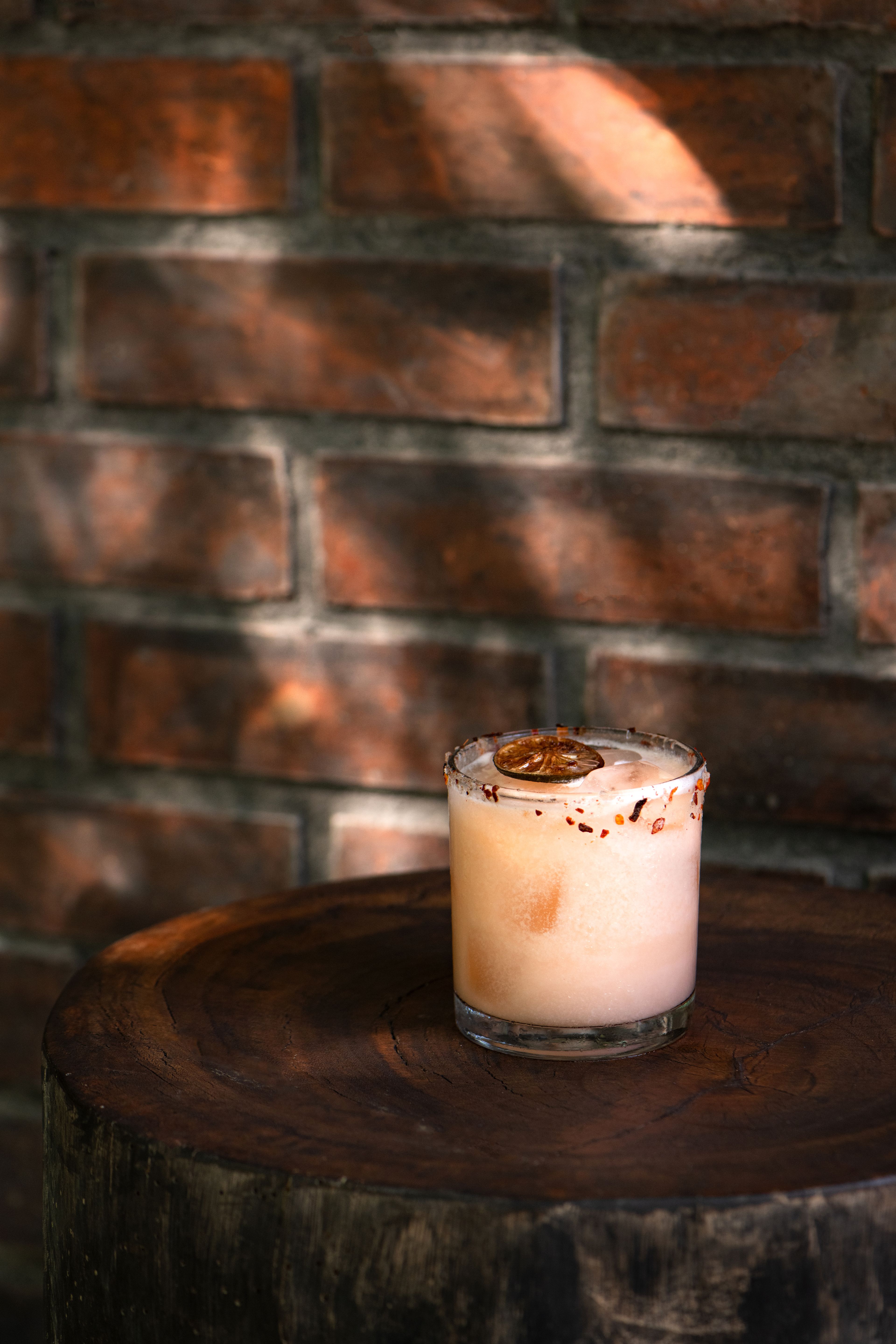 A pale cocktail with a dried citrus wheel on a rimmed glass, against a brick wall.