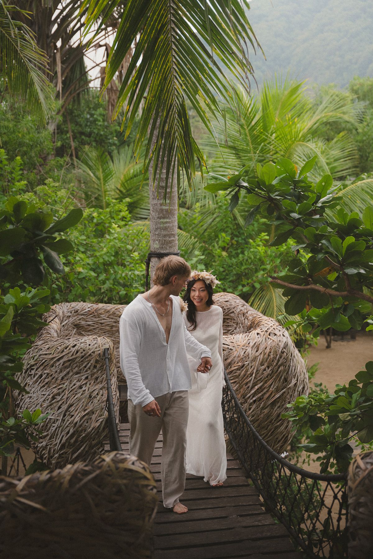 A couple in white standing together in the garden, the bride wearing a flower crown, woven pod chairs behind them.