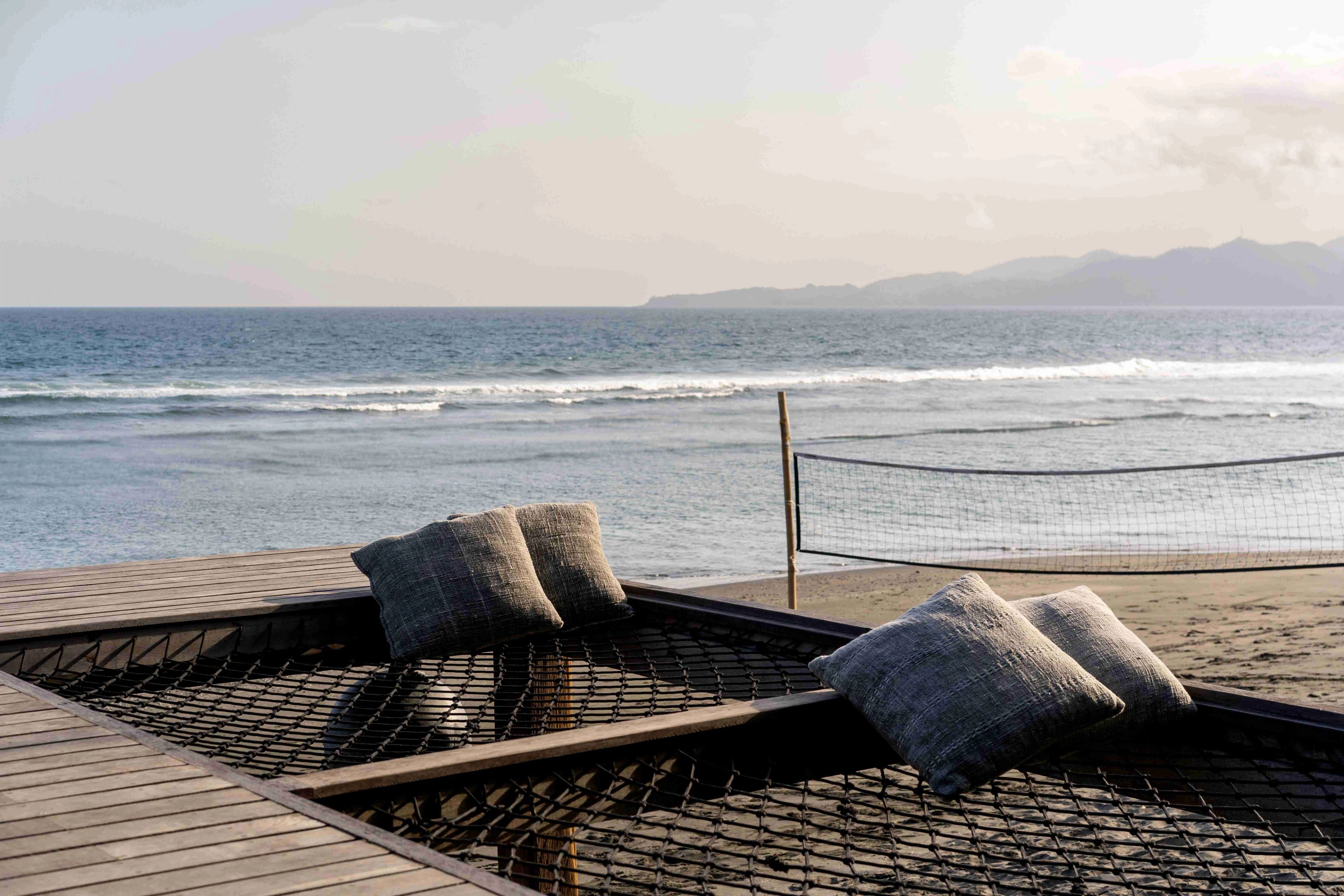 The rope-netted daybed and its cushions in flat early light, with the volleyball net on the sand and Nusa Penida across the water.