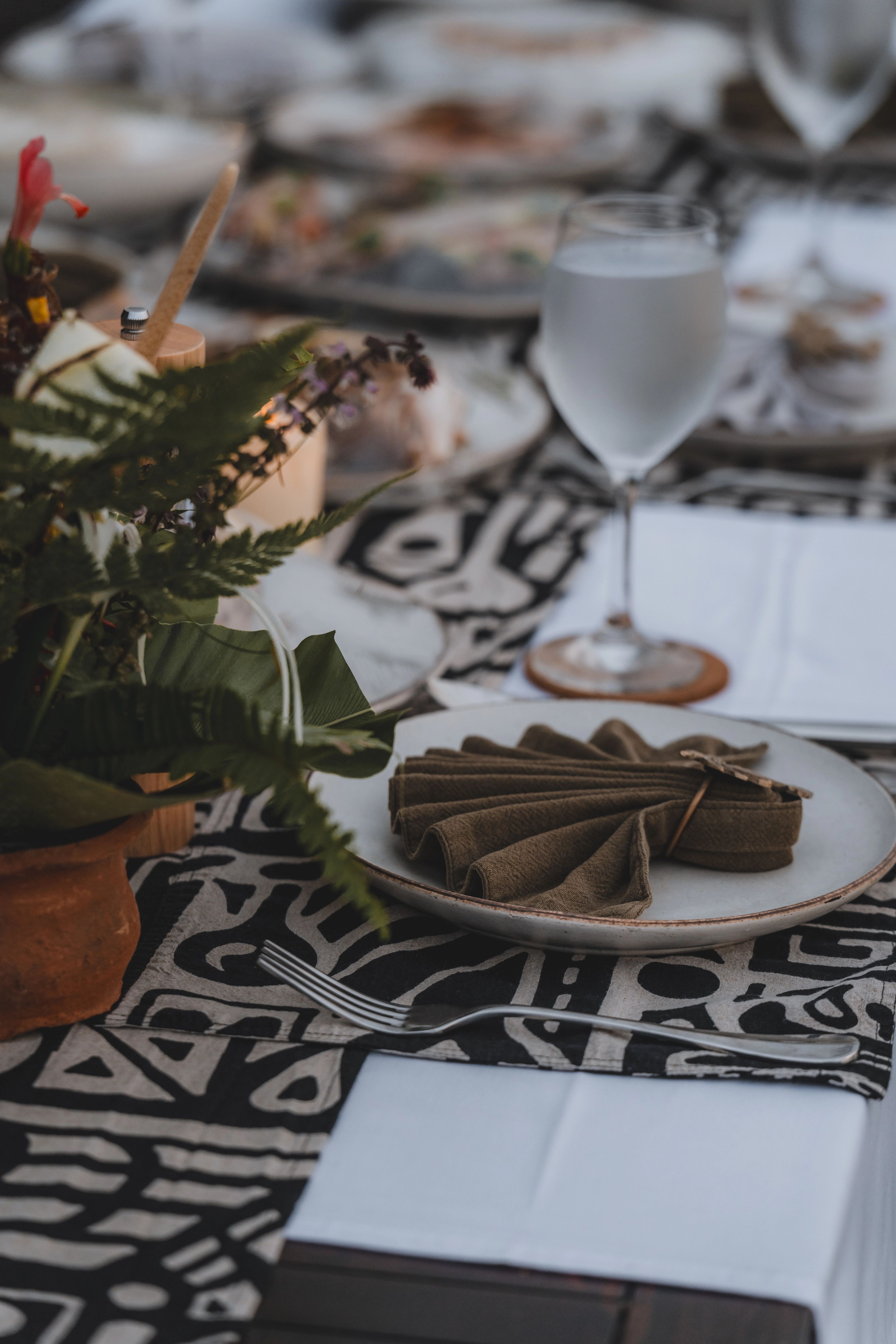 A folded olive napkin on a stoneware plate over a black-and-white patterned runner, with a wine glass and a fern arrangement beside it.
