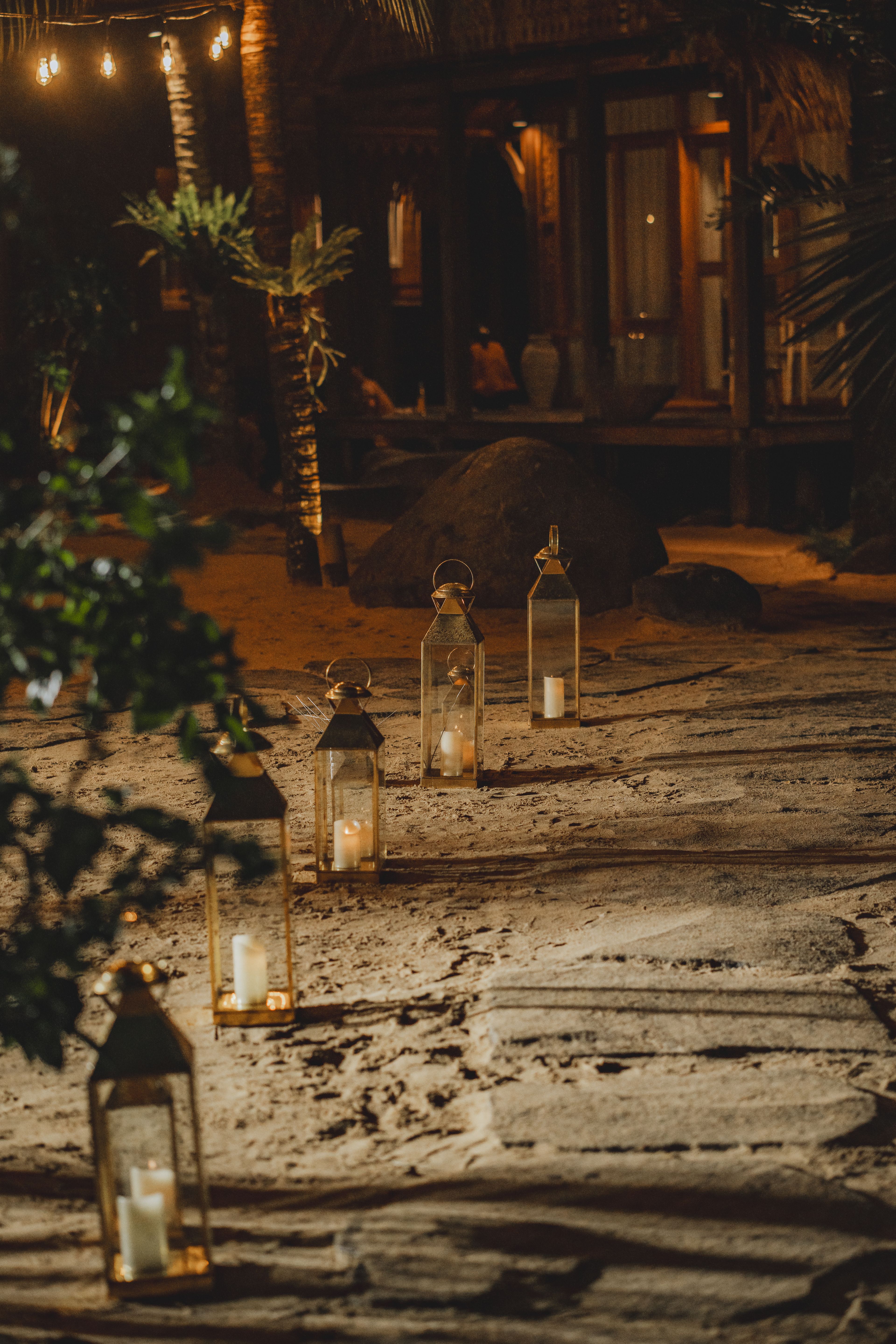 Glass hurricane lanterns lit along a path on the sand at night, seen past dark leaves.