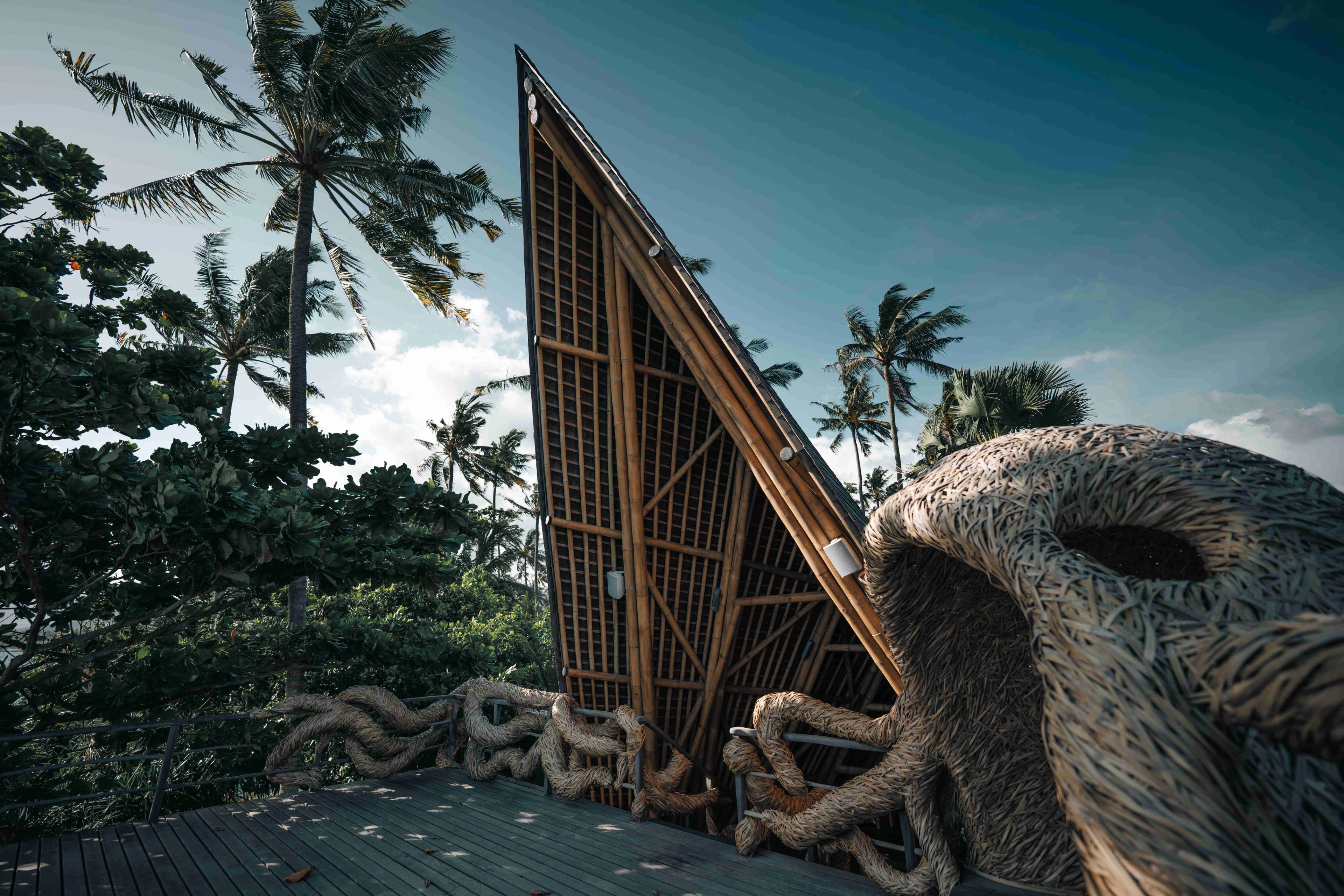 The pitched bamboo pavilion at the deck's edge, its woven rope sculpture set against palms and sky.