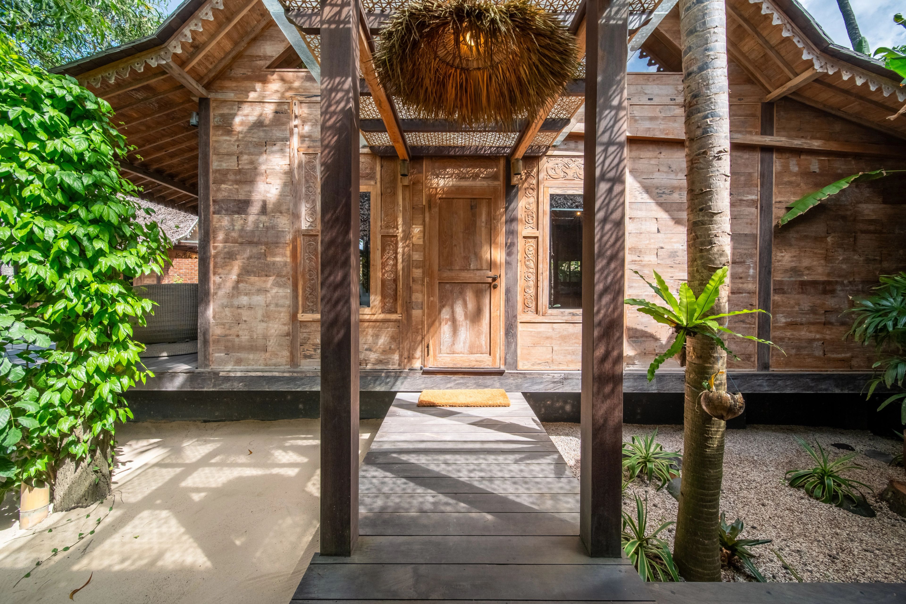 The villa's timber entrance framed by creeper and palms, a mat at the threshold.