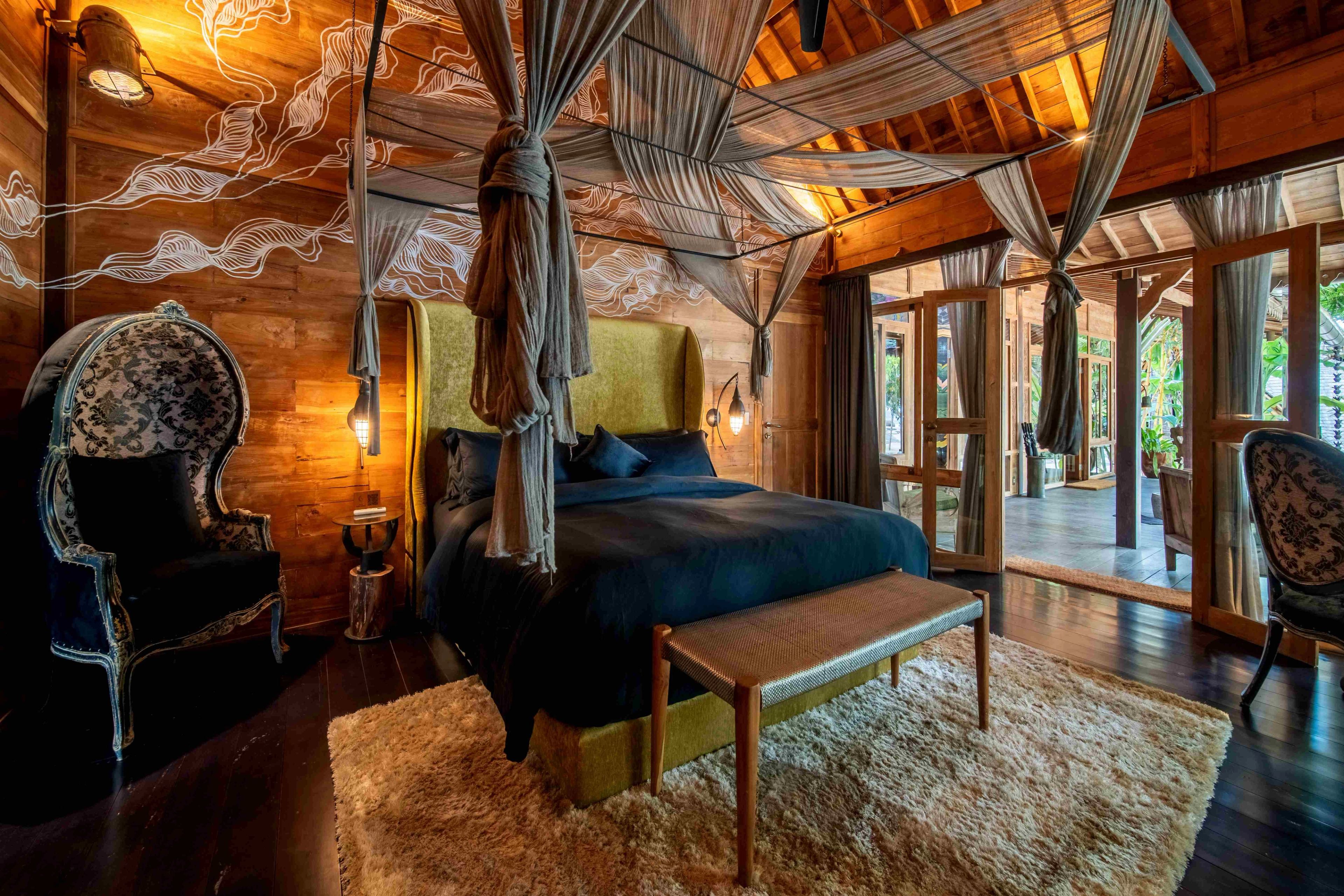 A bed under a draped canopy with carved chairs either side, doors open onto the deck.