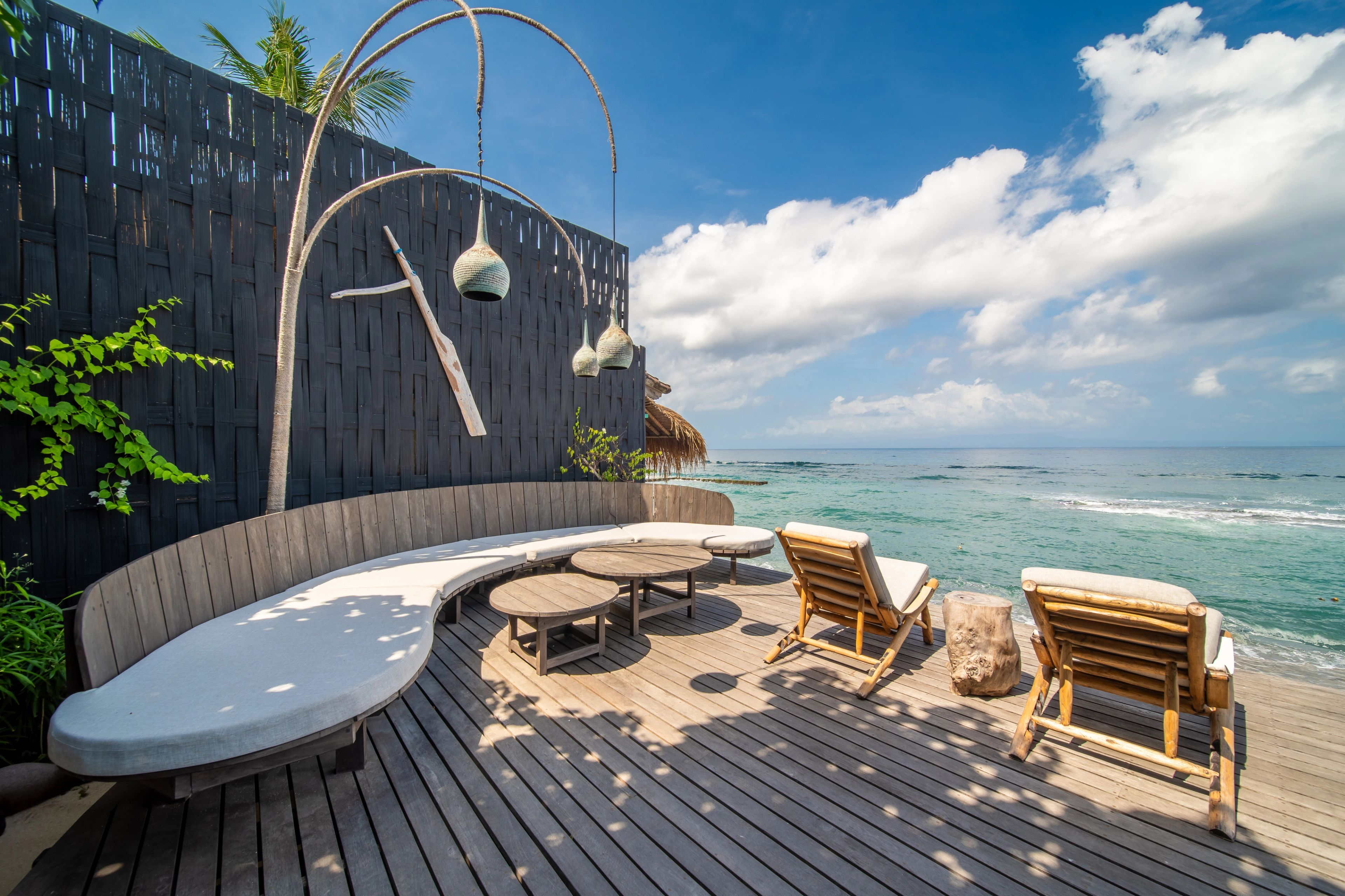A curved sofa lounge under woven pendant lamps, enclosed by a dark timber screen open to the ocean.