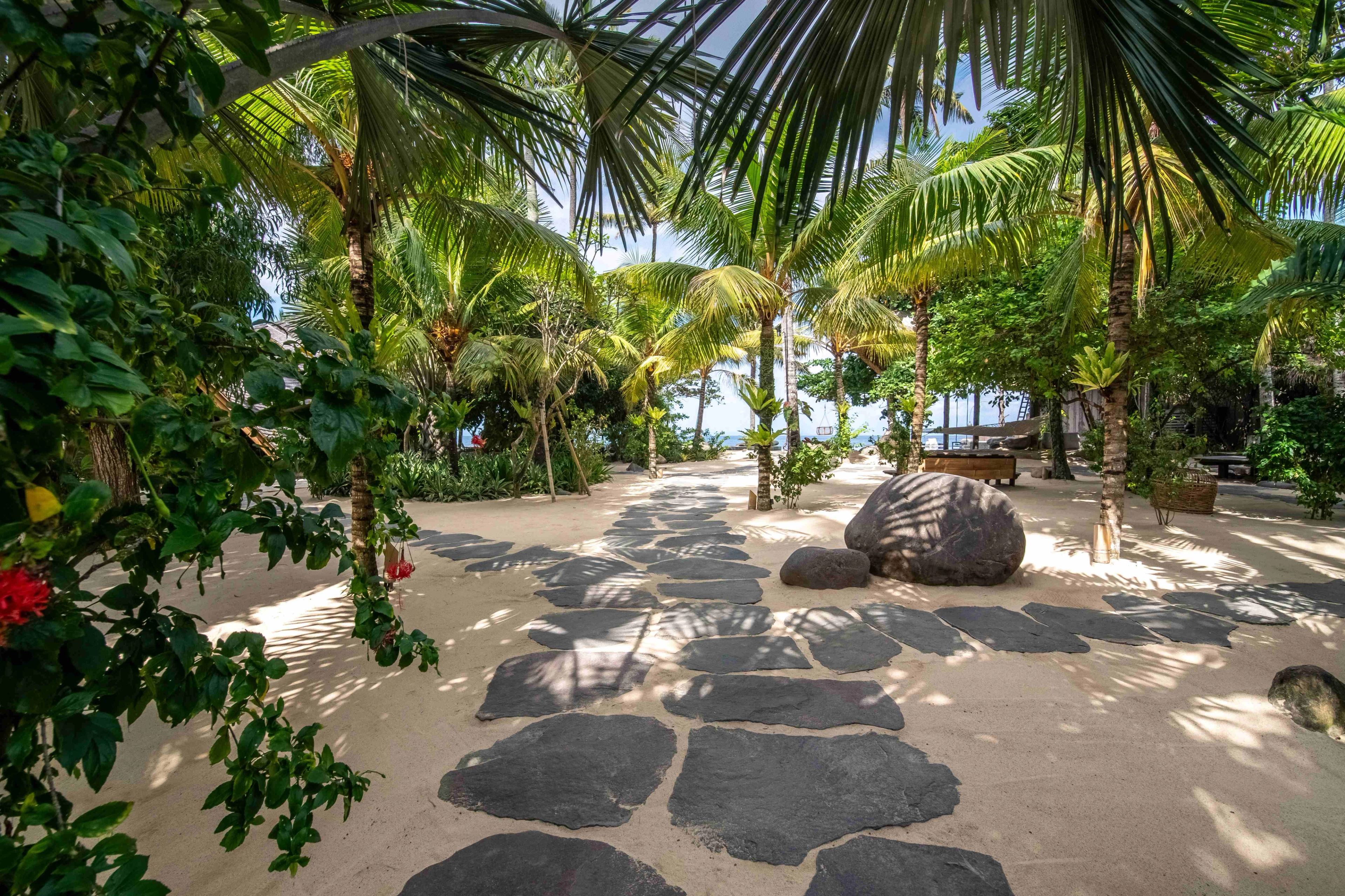 Stepping stones curving across the sand between palms, hibiscus flowering in the foreground.