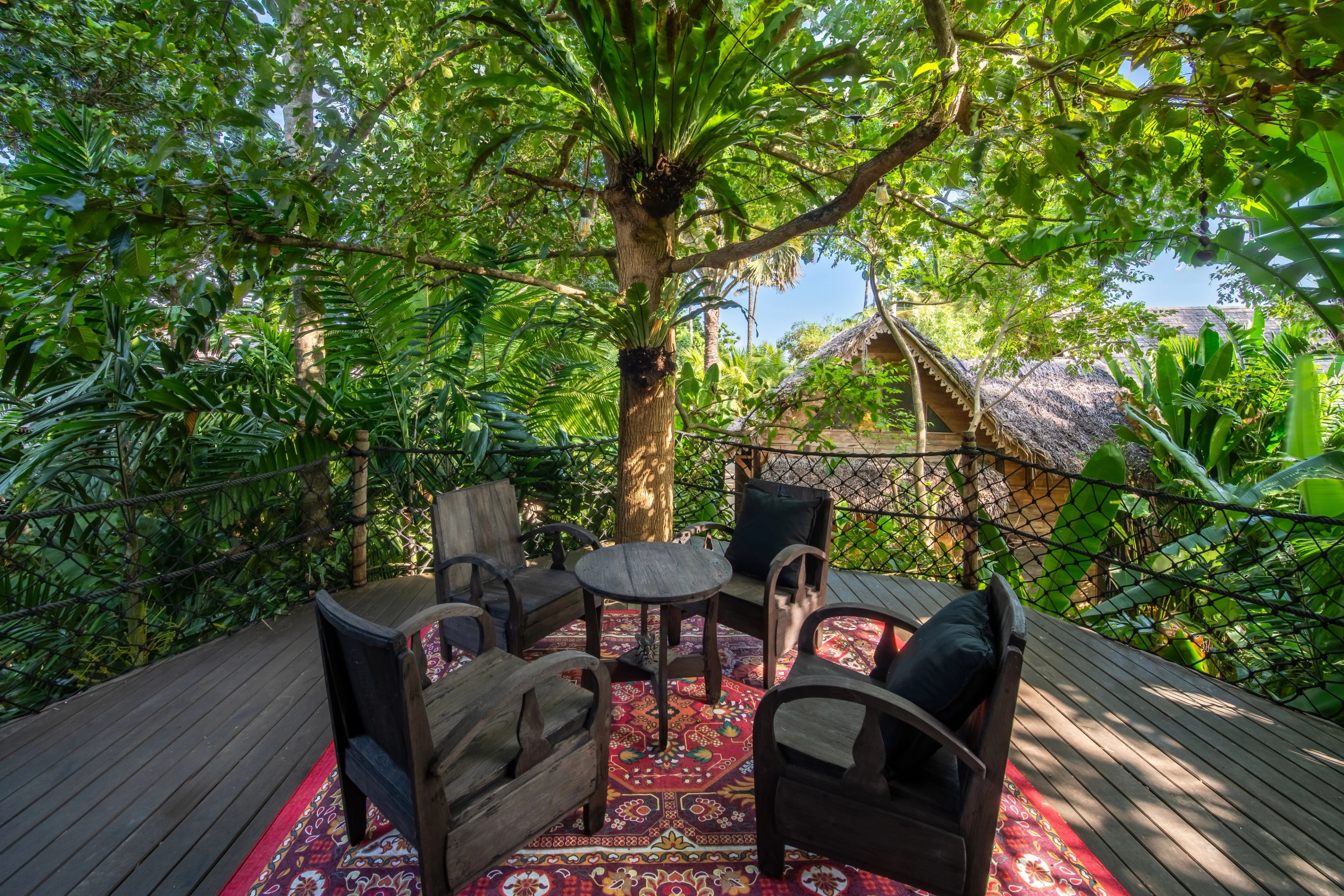 Timber armchairs and a low table on a patterned rug, on the deck under the trees.