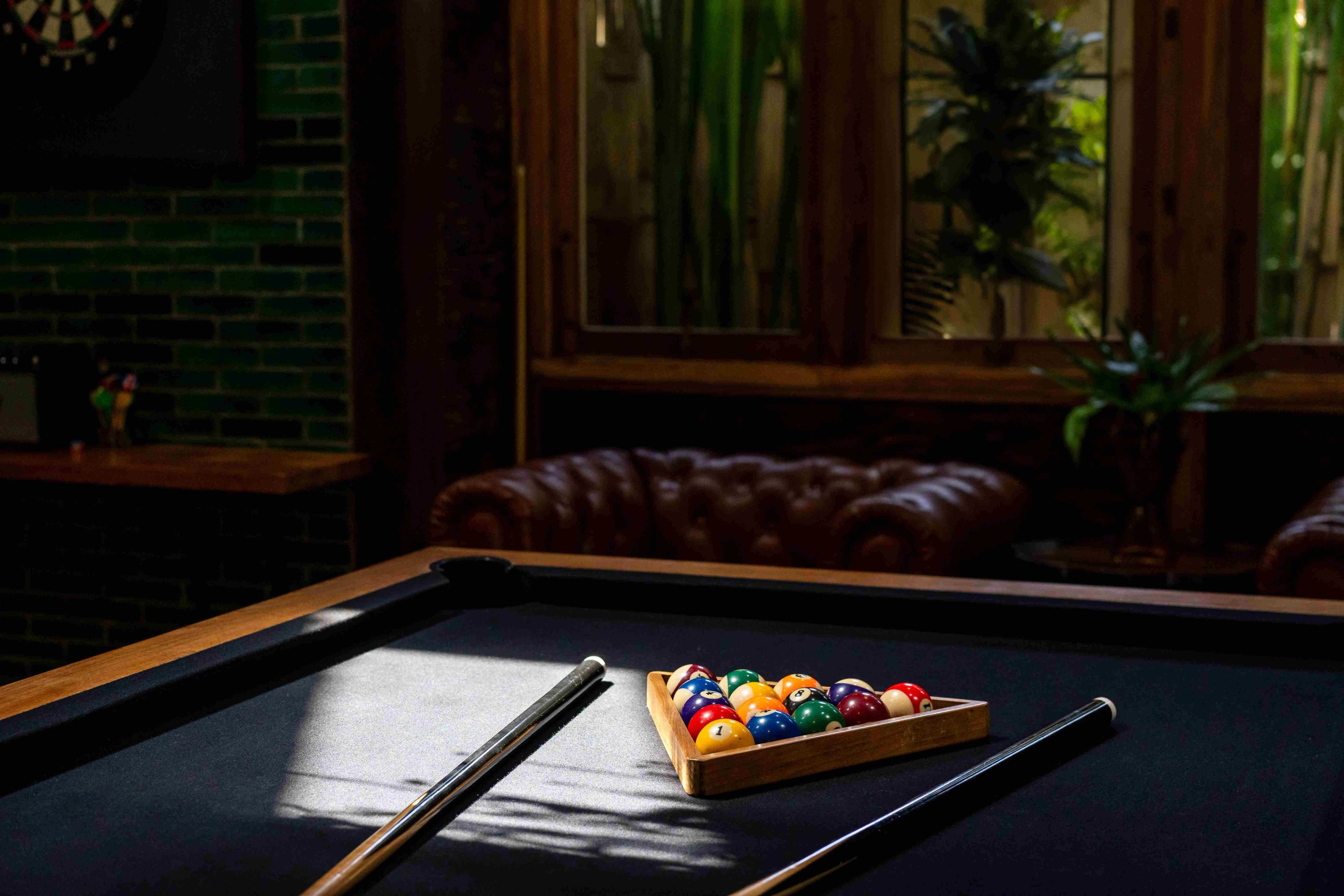 Racked balls and cues on the pool table, a leather sofa and dartboard behind it.