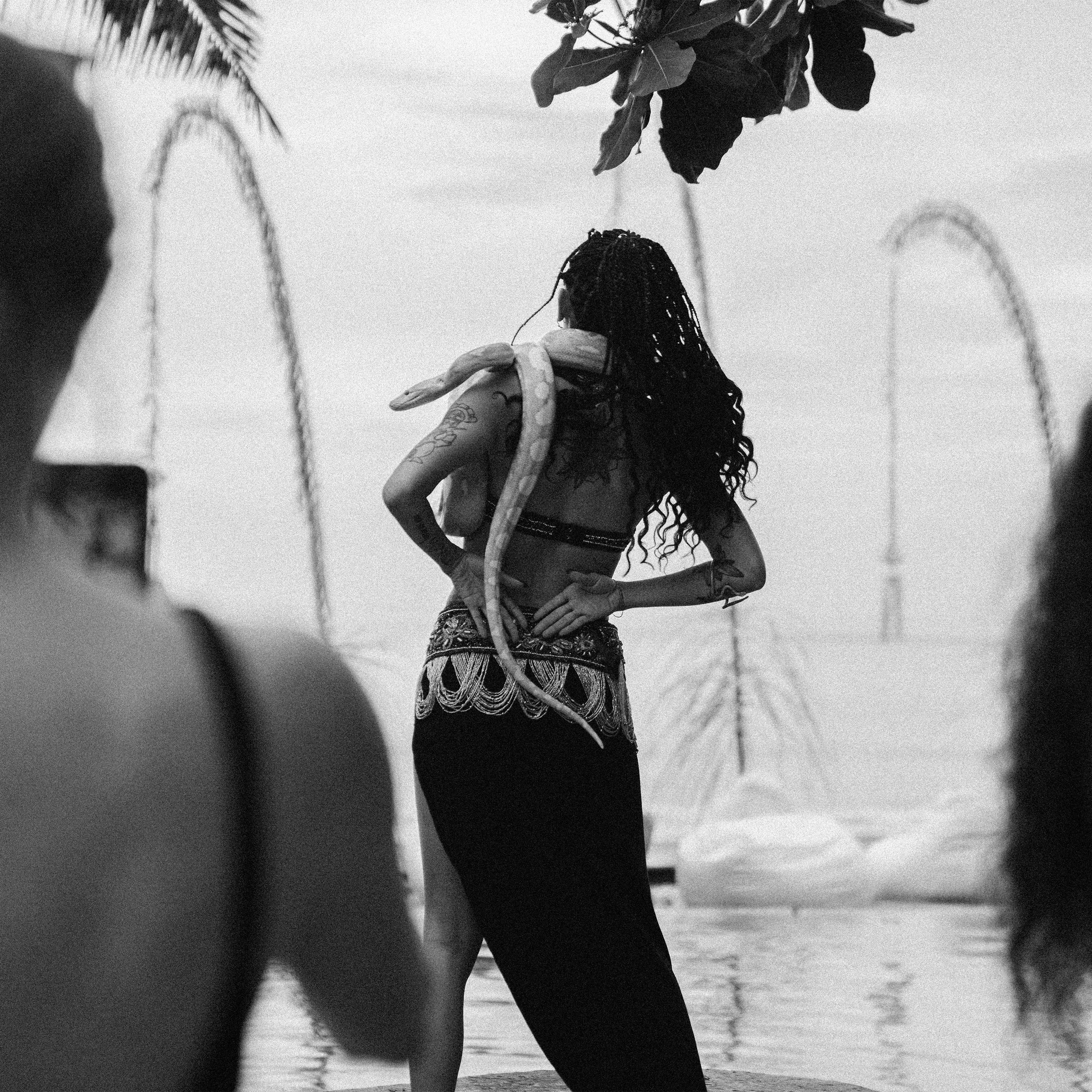 A black-and-white photograph of a dancer with a snake draped across her shoulders, fire jets either side.