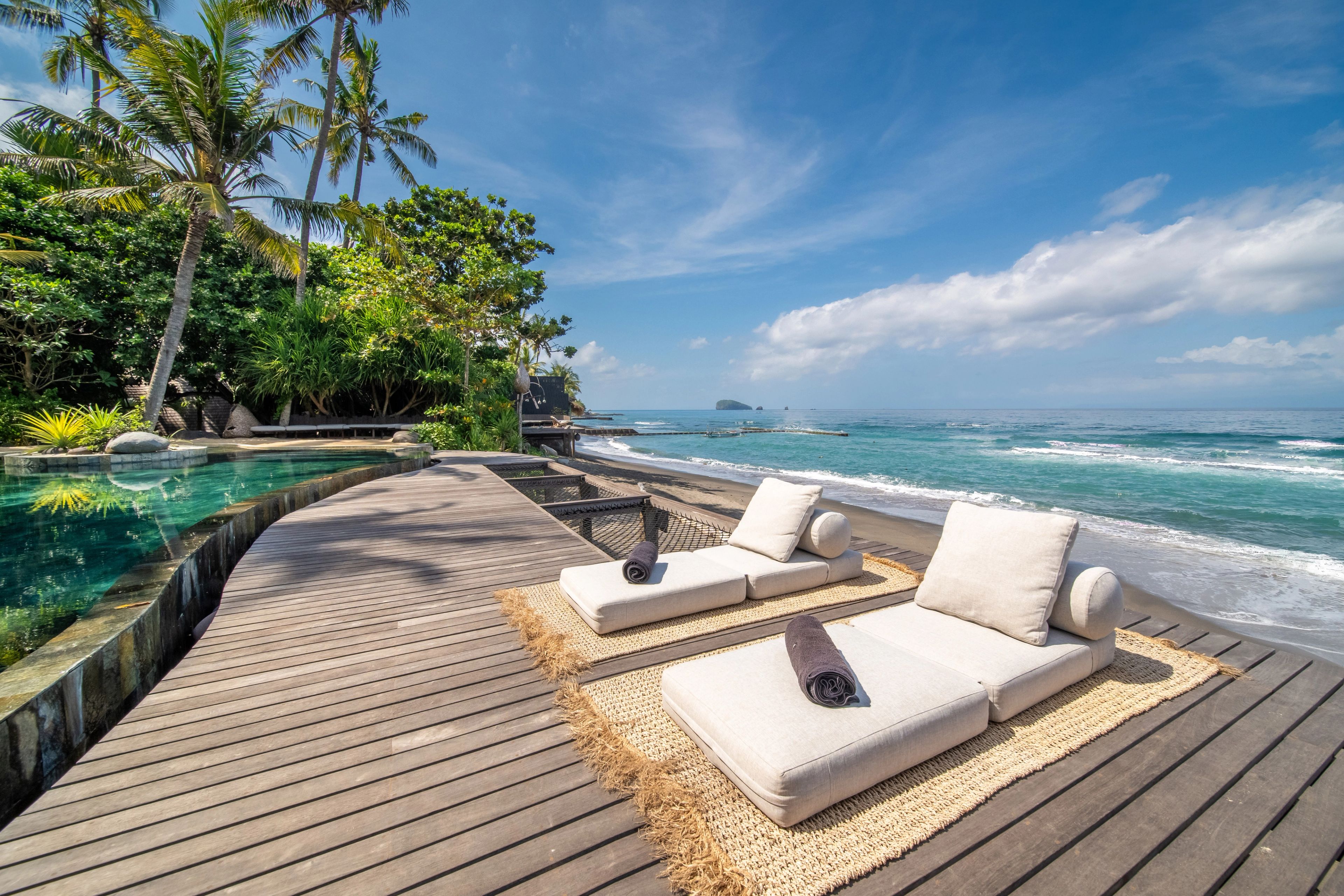 Two daybeds on a woven mat at the timber deck's edge, the pool on one side and the sea ahead.