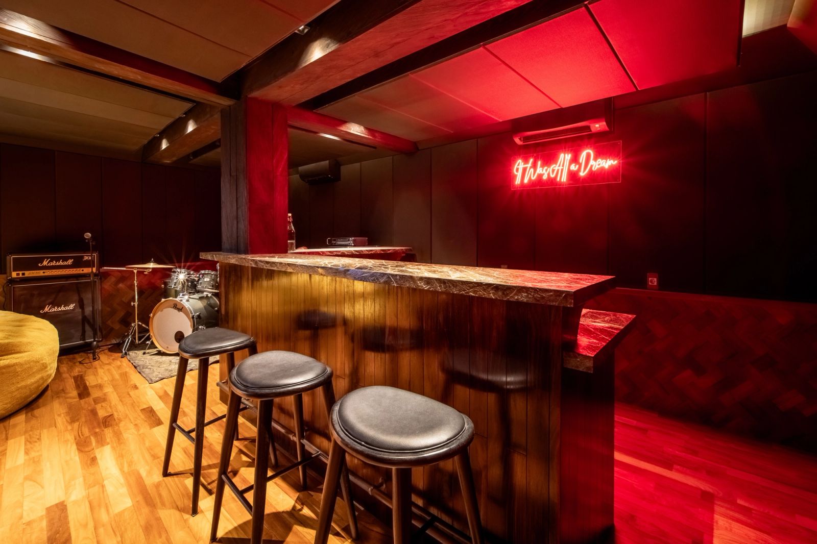 Stools along the timber bar under red neon, with the drum kit and amplifiers behind.
