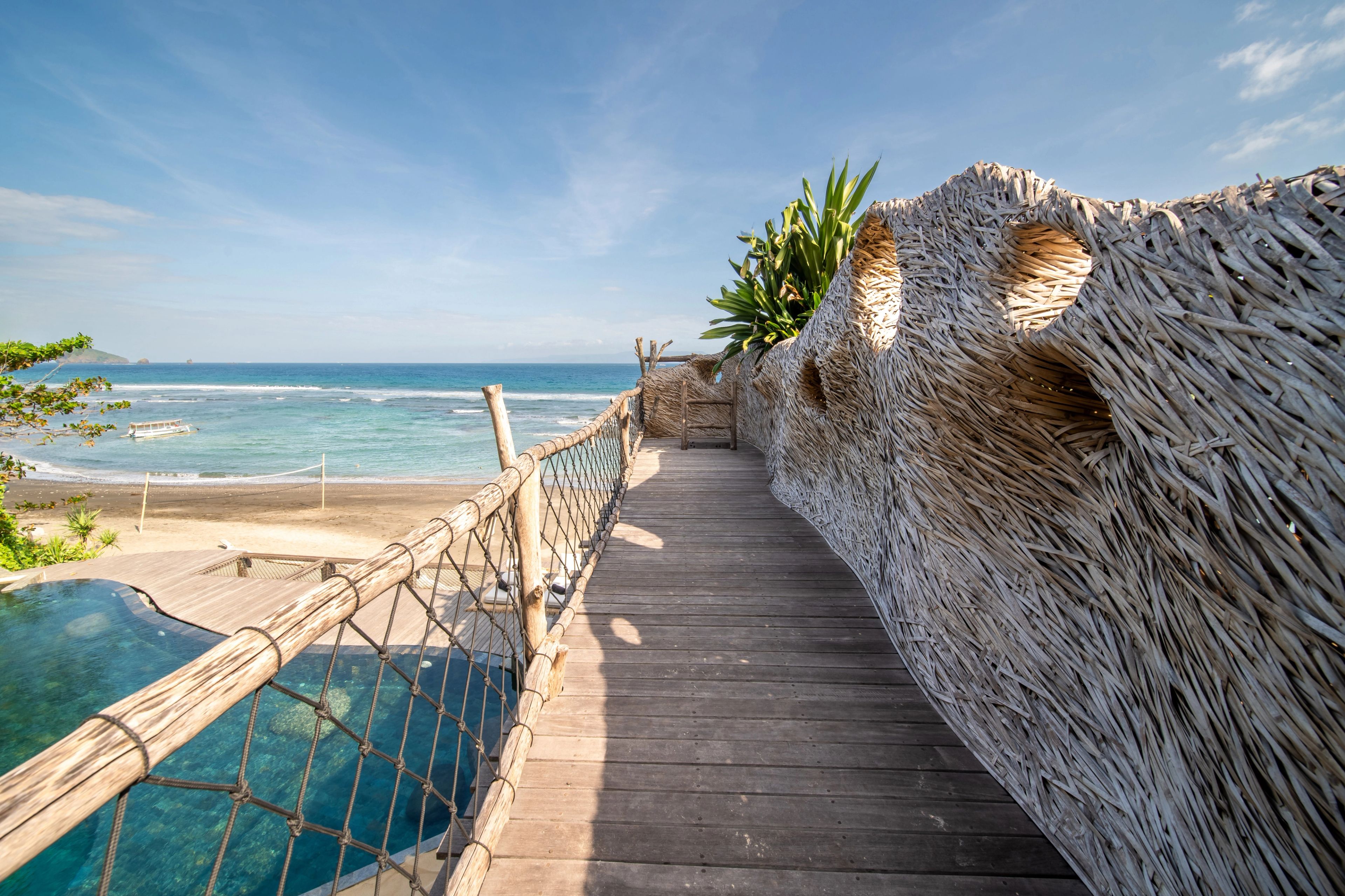 A rope-railed timber bridge running past a thatched woven pod towards the sand.