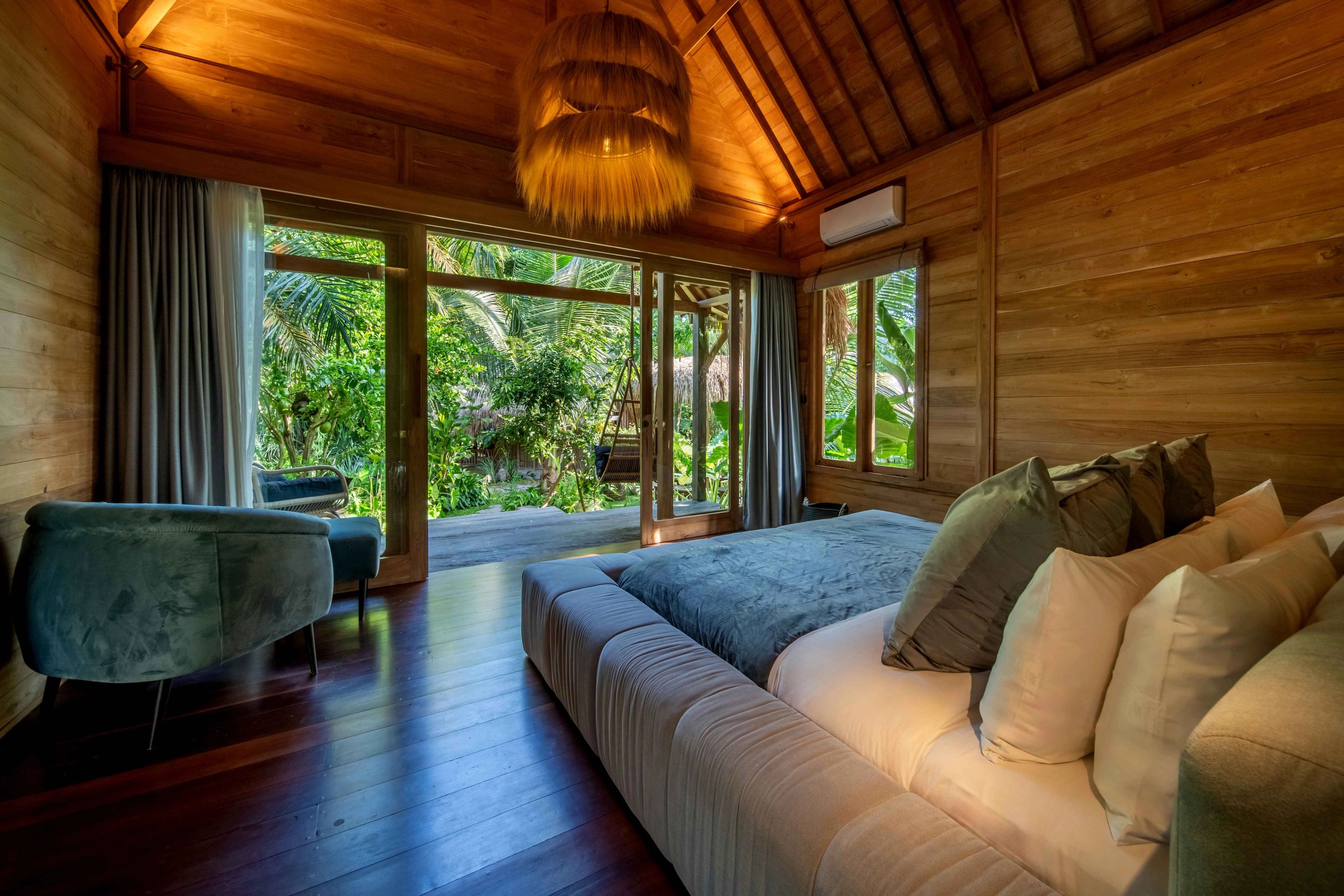 A bed piled with olive and cream cushions under a woven pendant, windows to the garden on two sides.